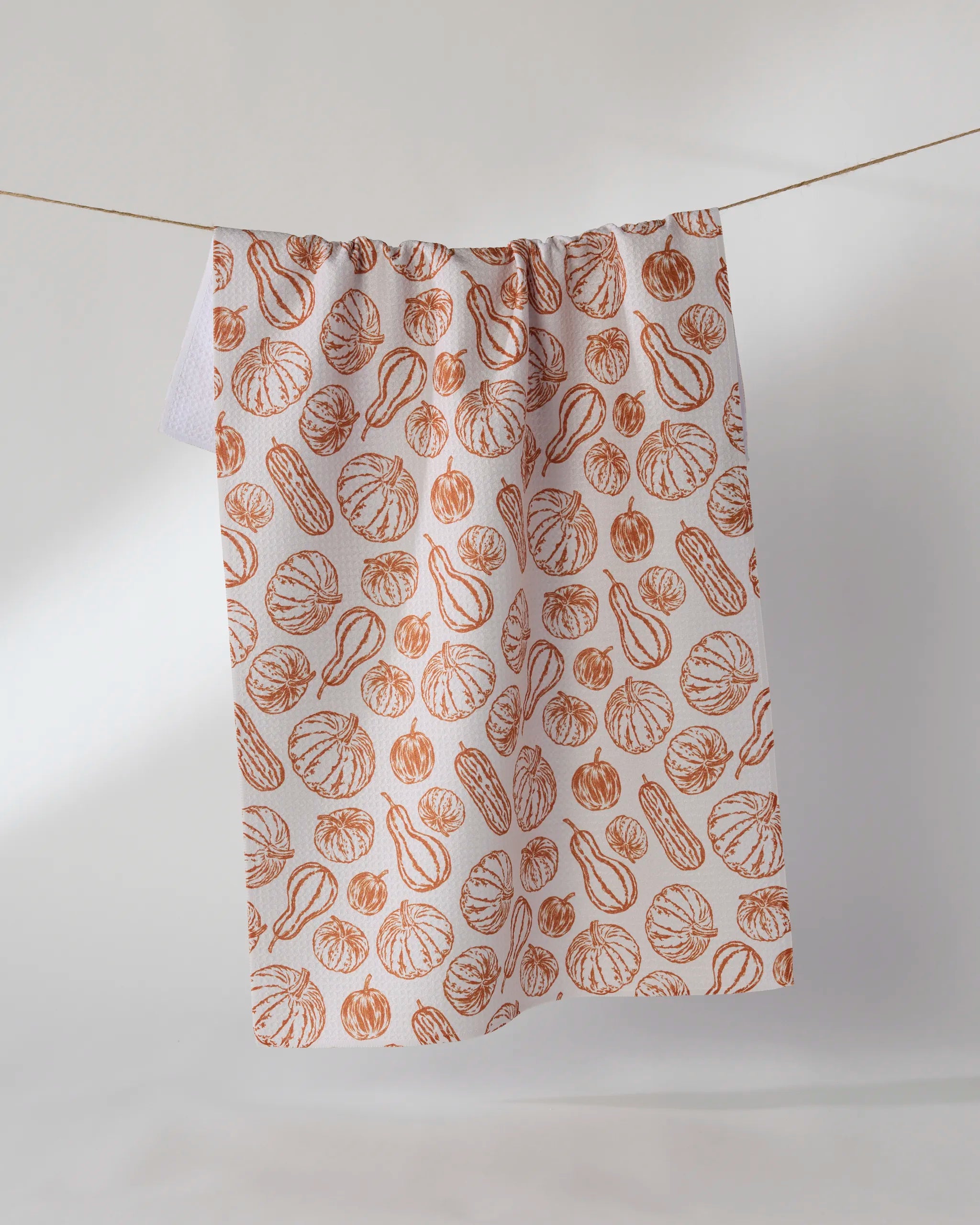 The Geometry Harvest Sketch tea towel features orange illustrations of assorted gourds and pumpkins on white, displayed hanging on a string against a plain white background.