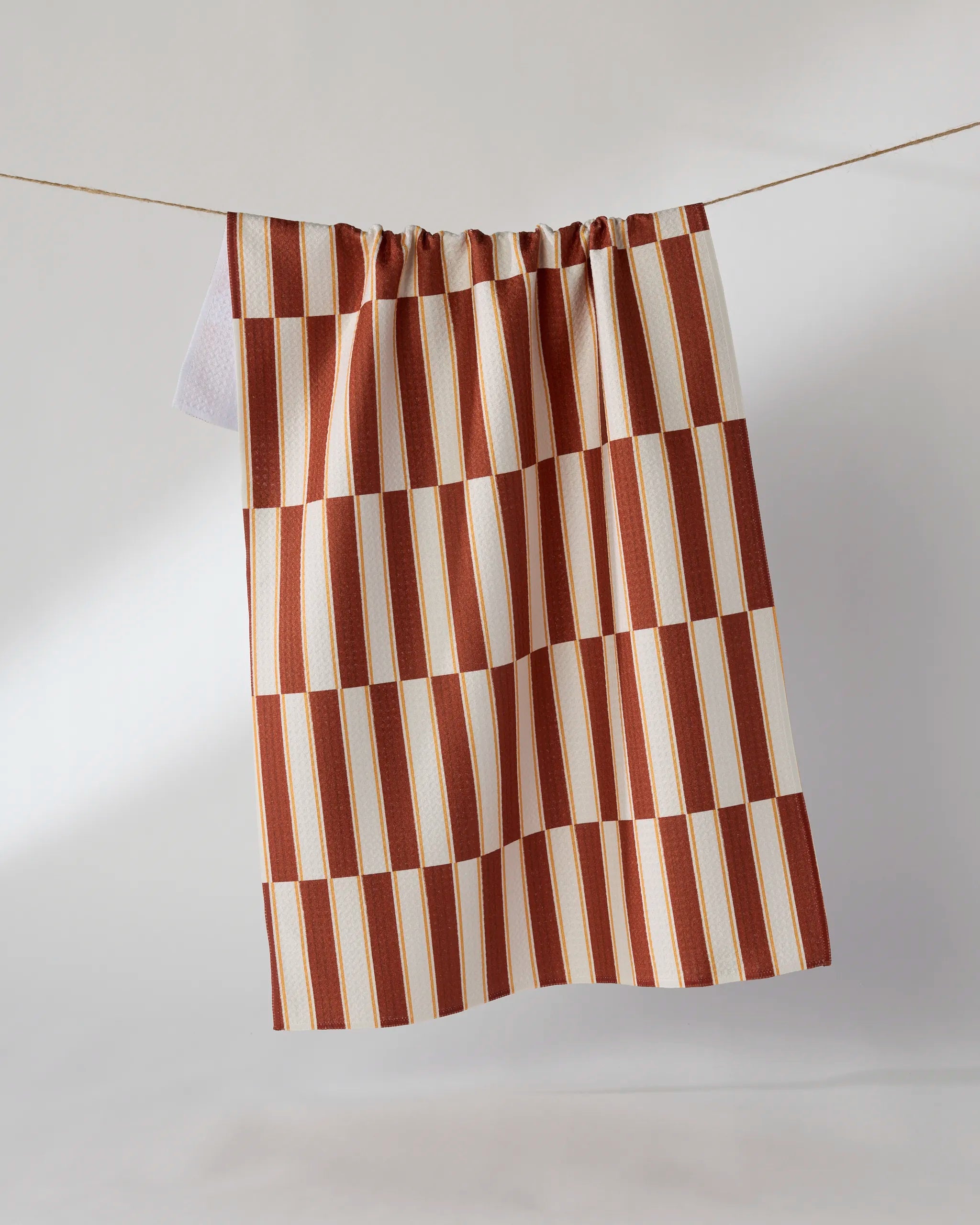 The Geometry Harvest Stripe tea towel features a red, beige, and white checkered and striped design and hangs on a clothesline against a plain white background. Made from recycled materials, it offers eco-friendly style for your kitchen.
