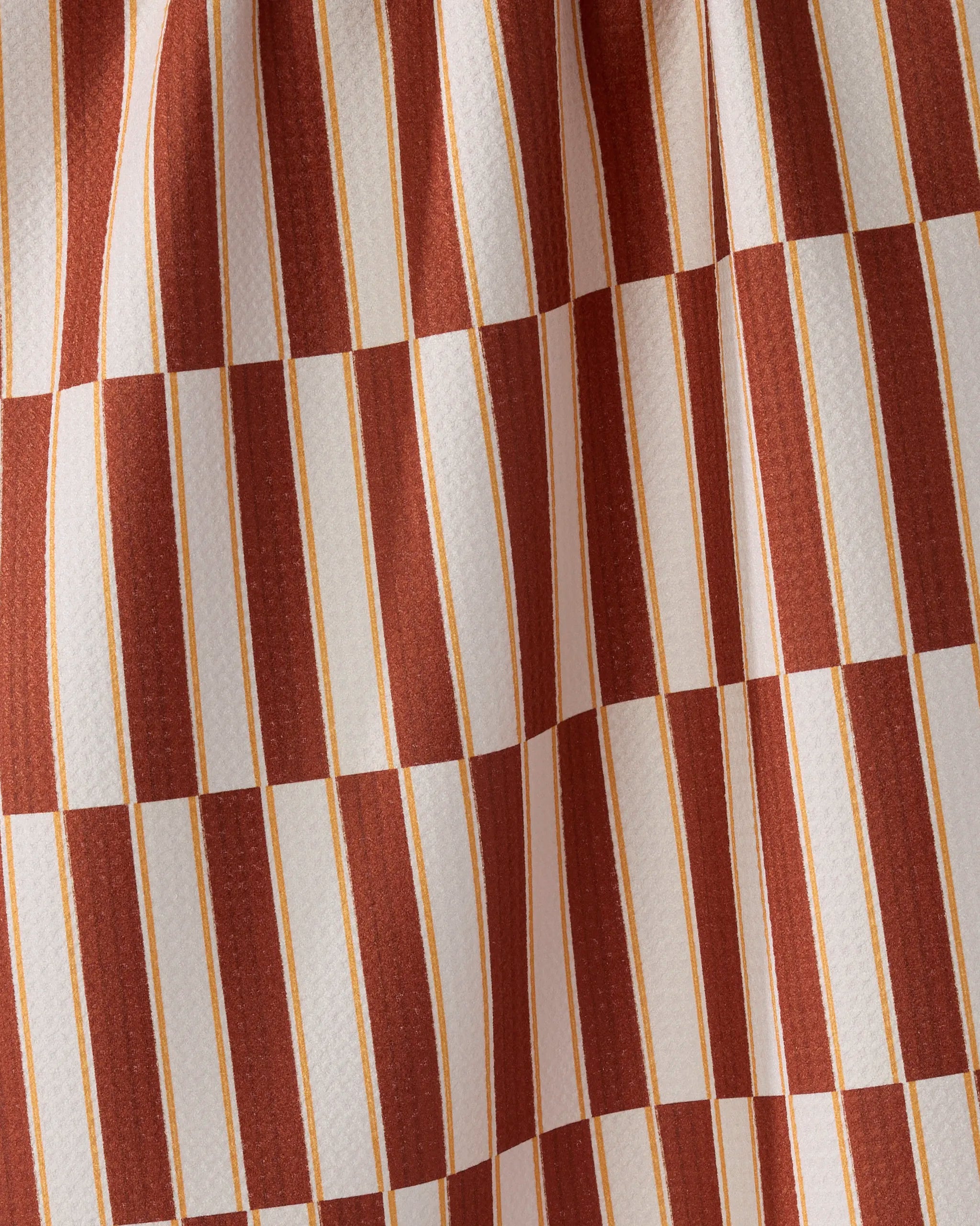 A close-up of the Geometry Harvest Stripe kitchen tea towel, featuring alternating brown and white stripes and checkerboard squares. Made from recycled materials, it has a textured surface with soft folds.