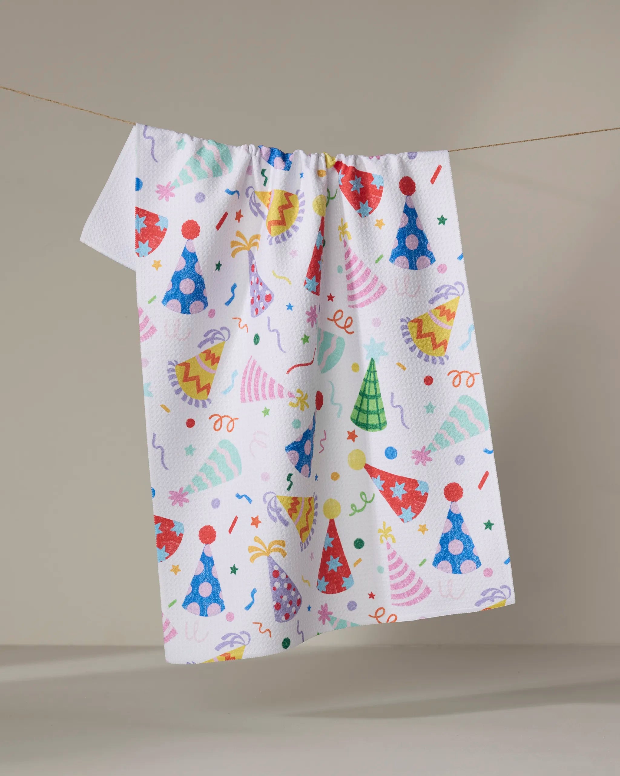 The "Hats Off" towel by Vaishali Design Studio, featuring colorful party hats, confetti, and streamers on a white ultra-absorbent fabric, hangs on a clothesline against a plain beige background.