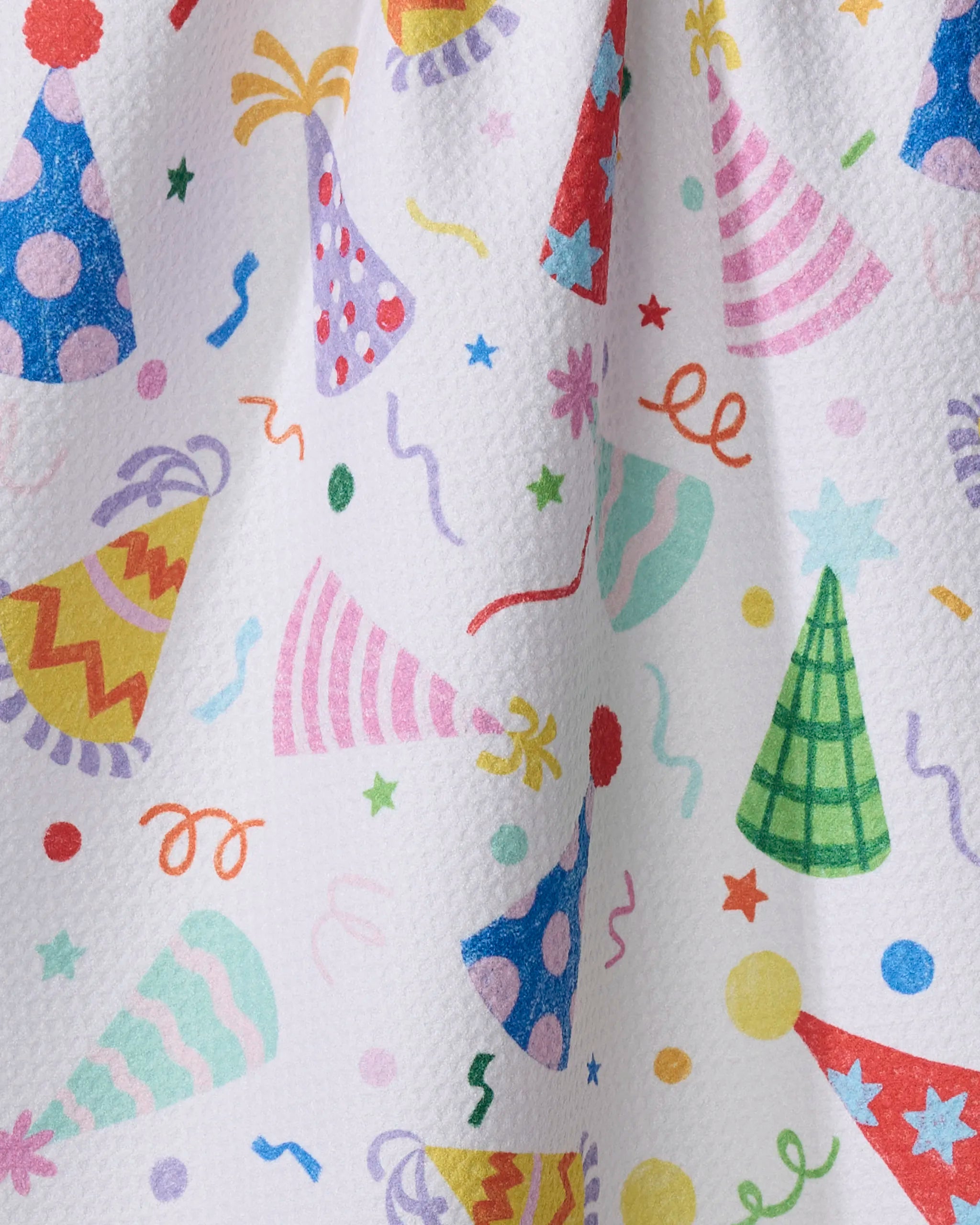 The "Hats Off" kitchen towel by Vaishali Design Studio features a festive celebration-themed design with colorful party hats, confetti, and streamers printed on textured white recycled fabric.