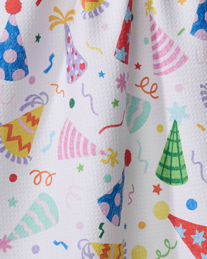 The "Hats Off" kitchen towel by Vaishali Design Studio features a festive celebration-themed design with colorful party hats, confetti, and streamers printed on textured white recycled fabric.