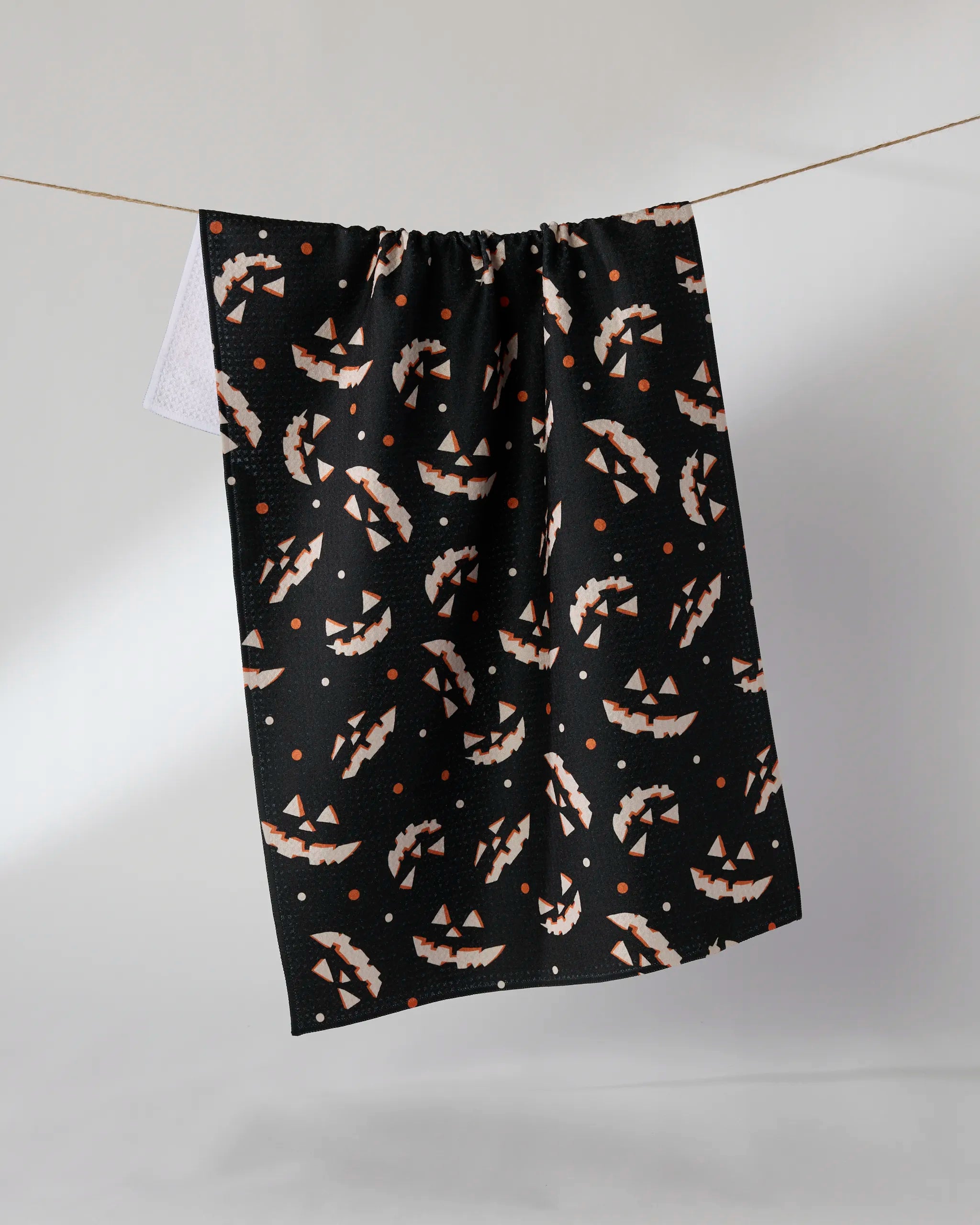The Geometry “Haunted Grin” black tea towel features an all-over pattern of orange and white jack-o’-lantern faces and small orange dots, hanging on a clothesline against a plain, light background.