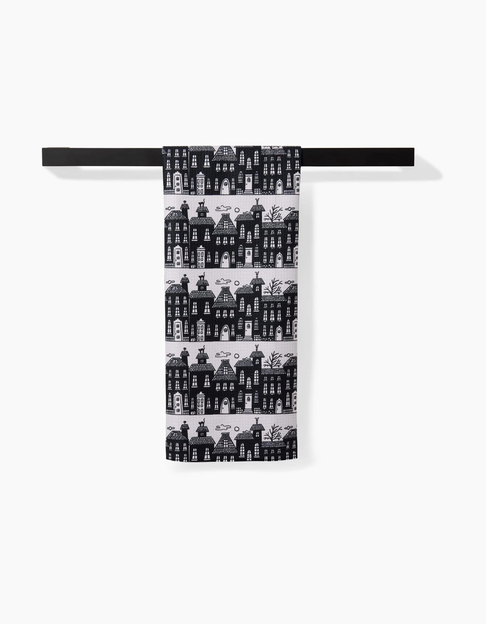 The Kayla Ann Haunted House Alley tea towel features a black and white pattern of illustrated houses, made from recycled materials, and hangs neatly on a minimalist black towel rack against a white background.