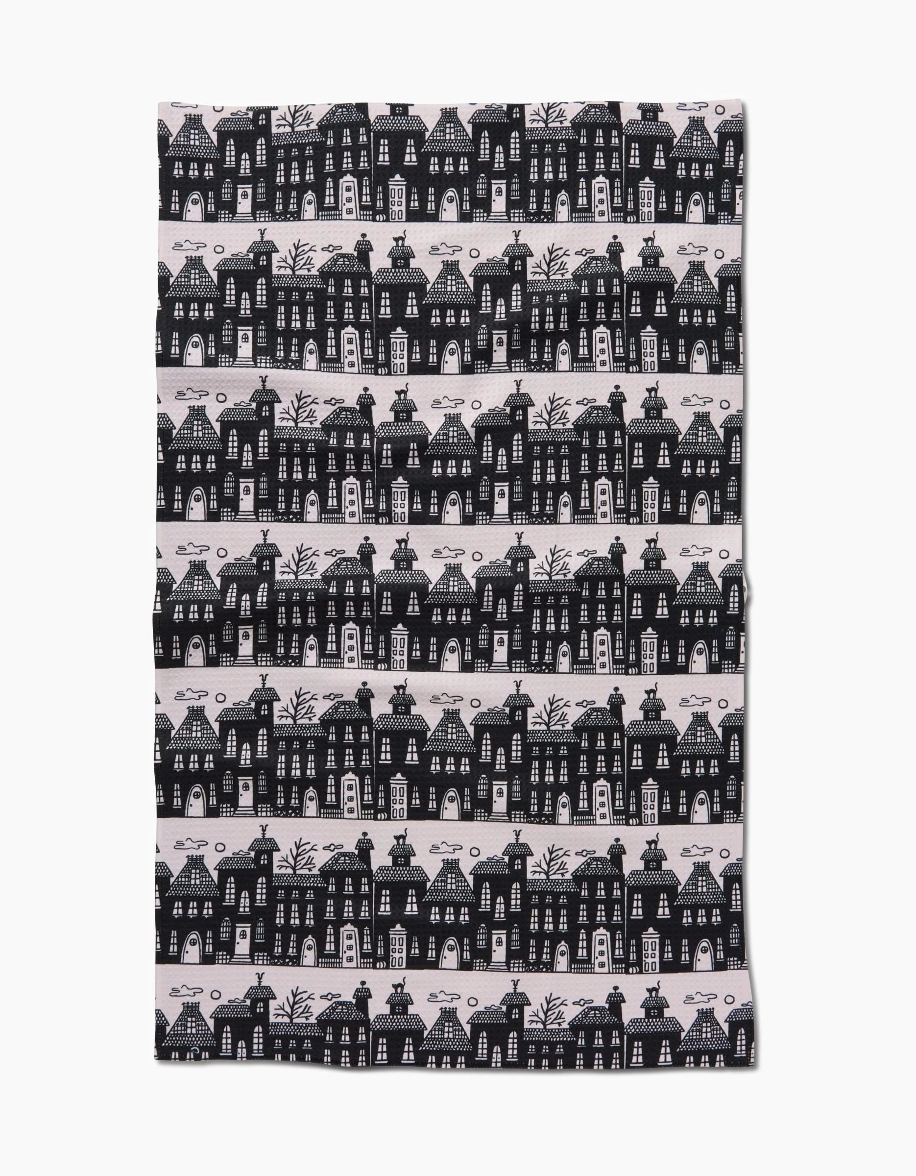 The Kayla Ann Haunted House Alley tea towel features whimsical black-and-white houses, trees, and clouds in horizontal rows on a light background. Made from recycled materials, its dark illustrations create a striking contrast.