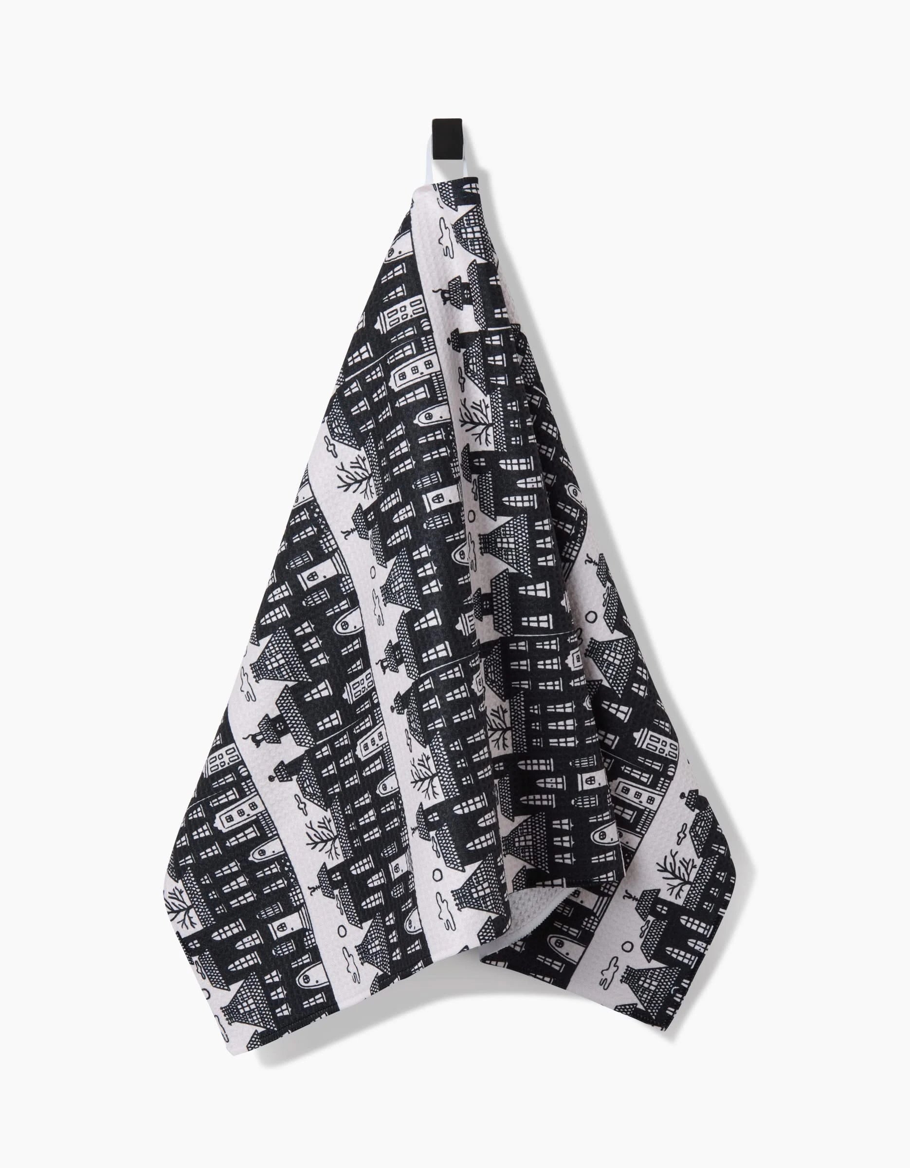 The Haunted House Alley tea towel by Kayla Ann, featuring a black and white pattern of buildings and trees and made from recycled materials, hangs from a loop against a plain white background.