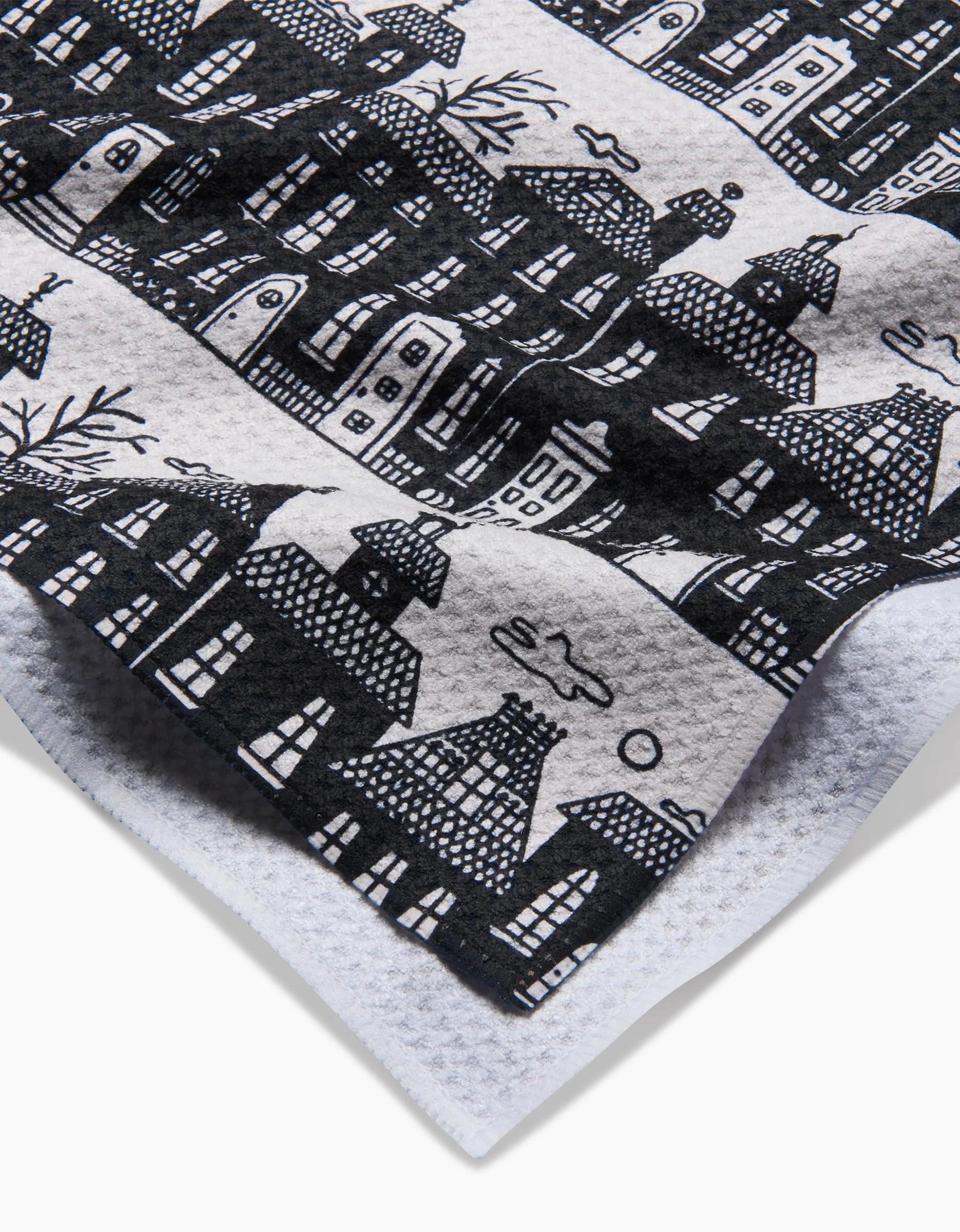 A close-up of the textured white Haunted House Alley tea towel by Kayla Ann, featuring a black hand-drawn print of whimsical houses, trees, clouds, and the sun. The ultra-absorbent cloth is playfully styled and slightly folded at the corner.