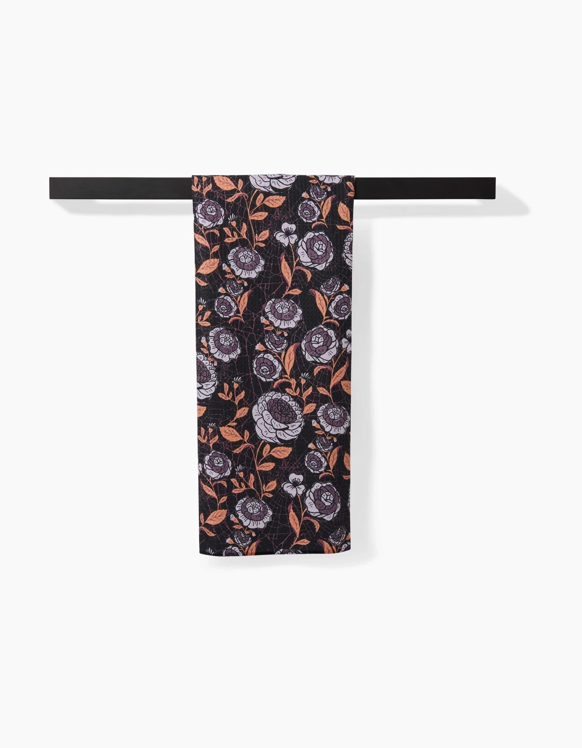 The Haunted Roses tea towel by Danelys Sidron, featuring a purple and orange floral pattern, hangs neatly over a horizontal black bar against a plain white background.