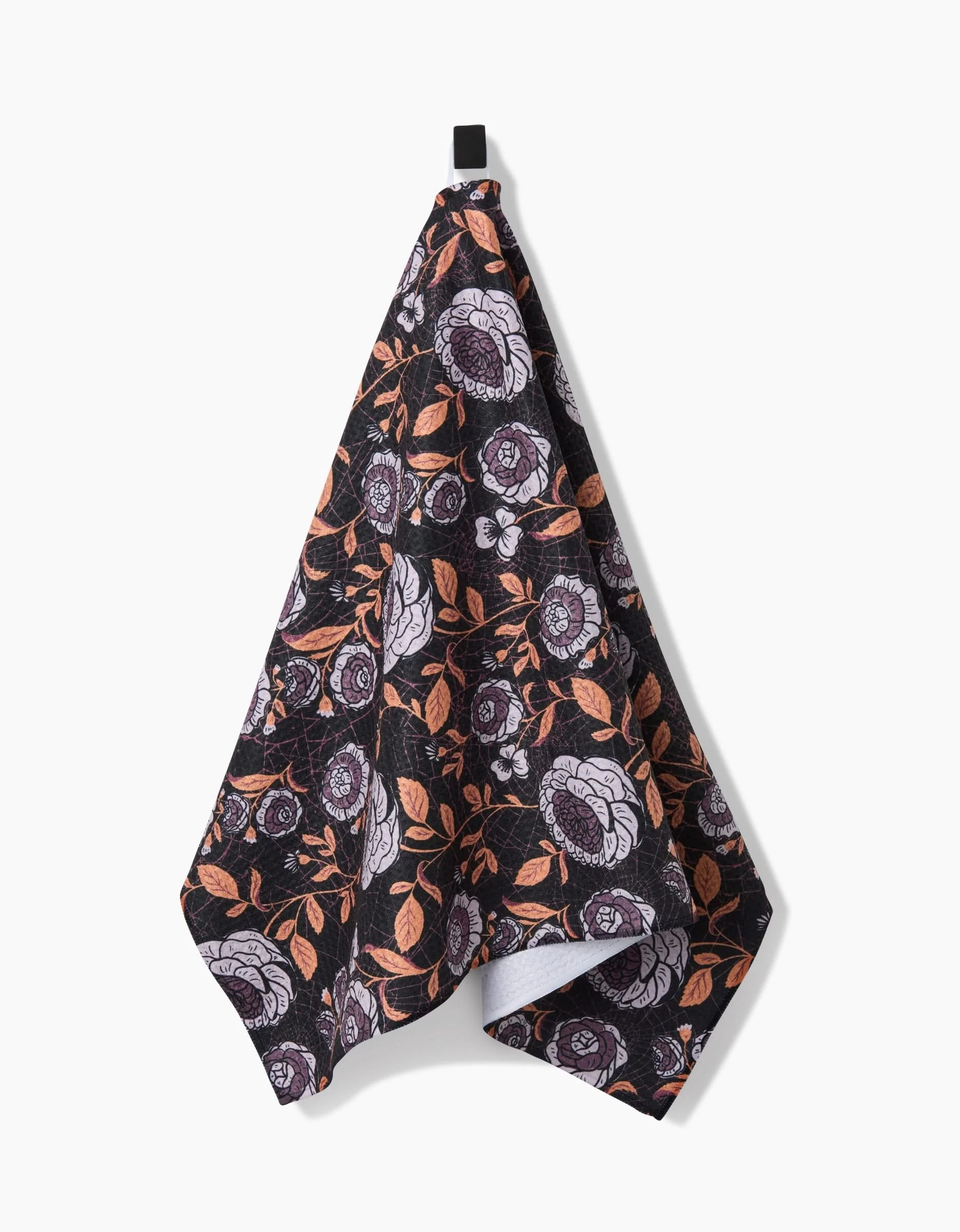 The Haunted Roses black kitchen towel by Danelys Sidron features a floral pattern with white and purple flowers and orange leaves, made from recycled materials, hanging on a black hook against a plain white background.