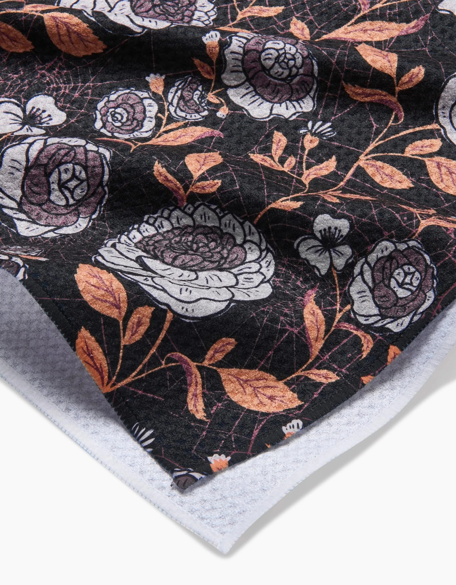 A close-up of the “Haunted Roses” kitchen towel by Danelys Sidron shows large white and pink flowers, orange leaves, and thin branches on a black background, with a textured white backing at the corner.