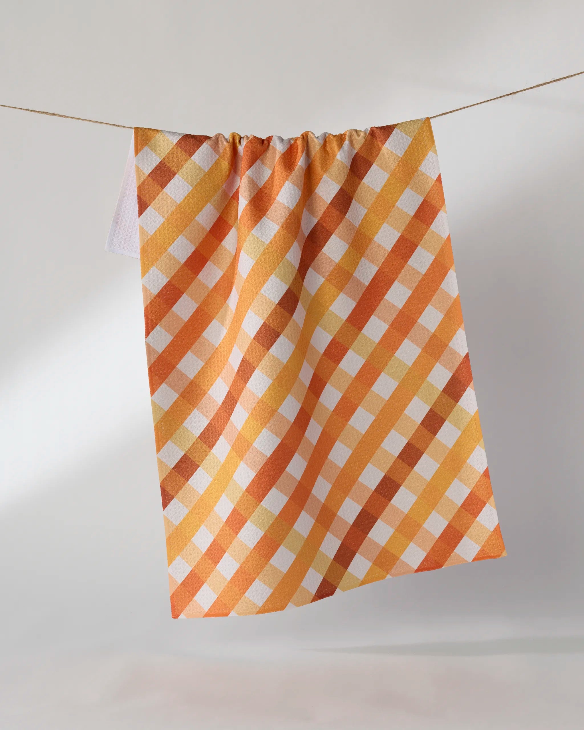 The Geometry Haybales recycled kitchen towel, featuring a checkered pattern in orange, yellow, and white, hangs on a clothesline against a plain light background.