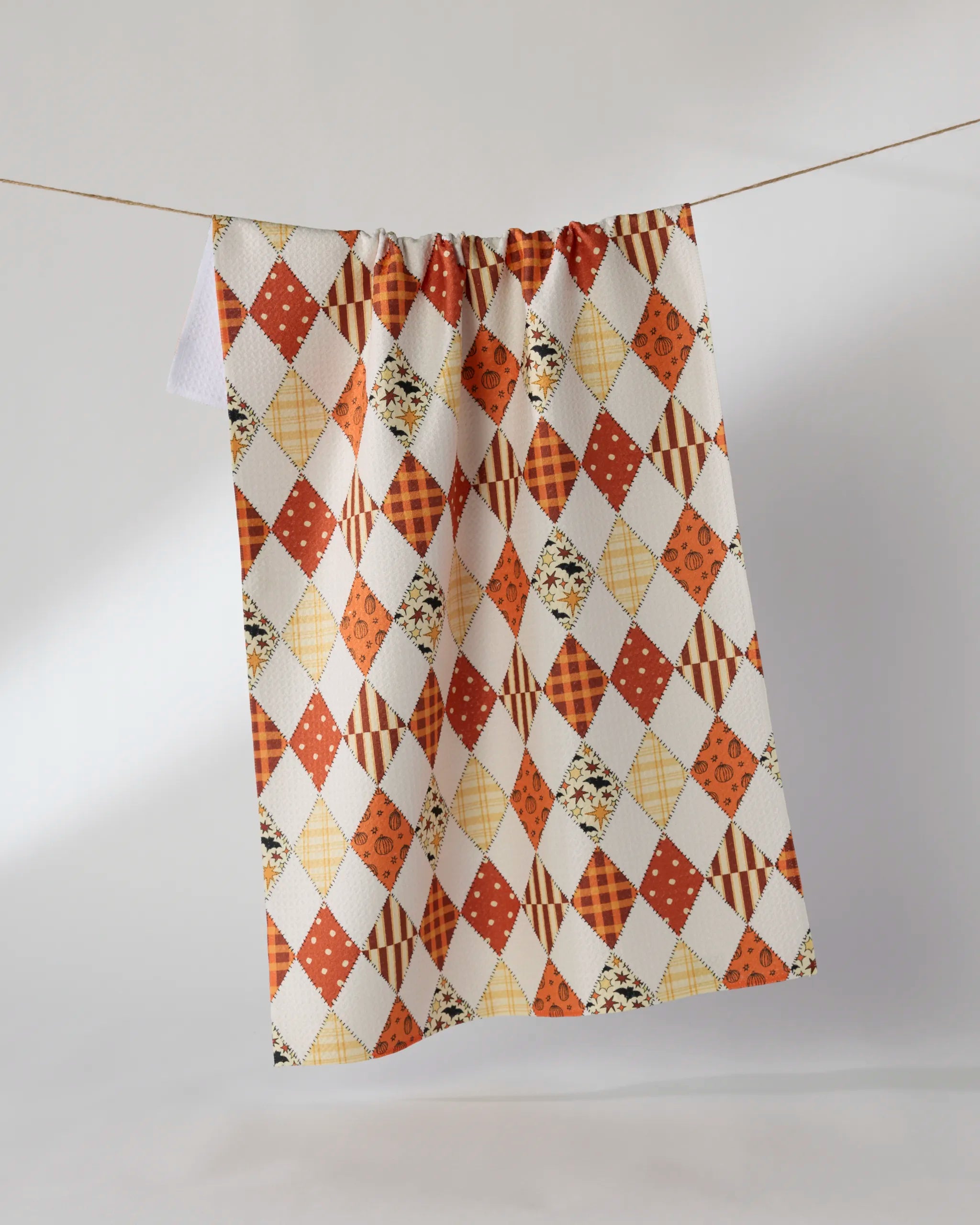 The Geometry Hayride recycled kitchen towel, featuring an orange, yellow, and brown patchwork diamond pattern, hangs on a clothesline against a plain white background.