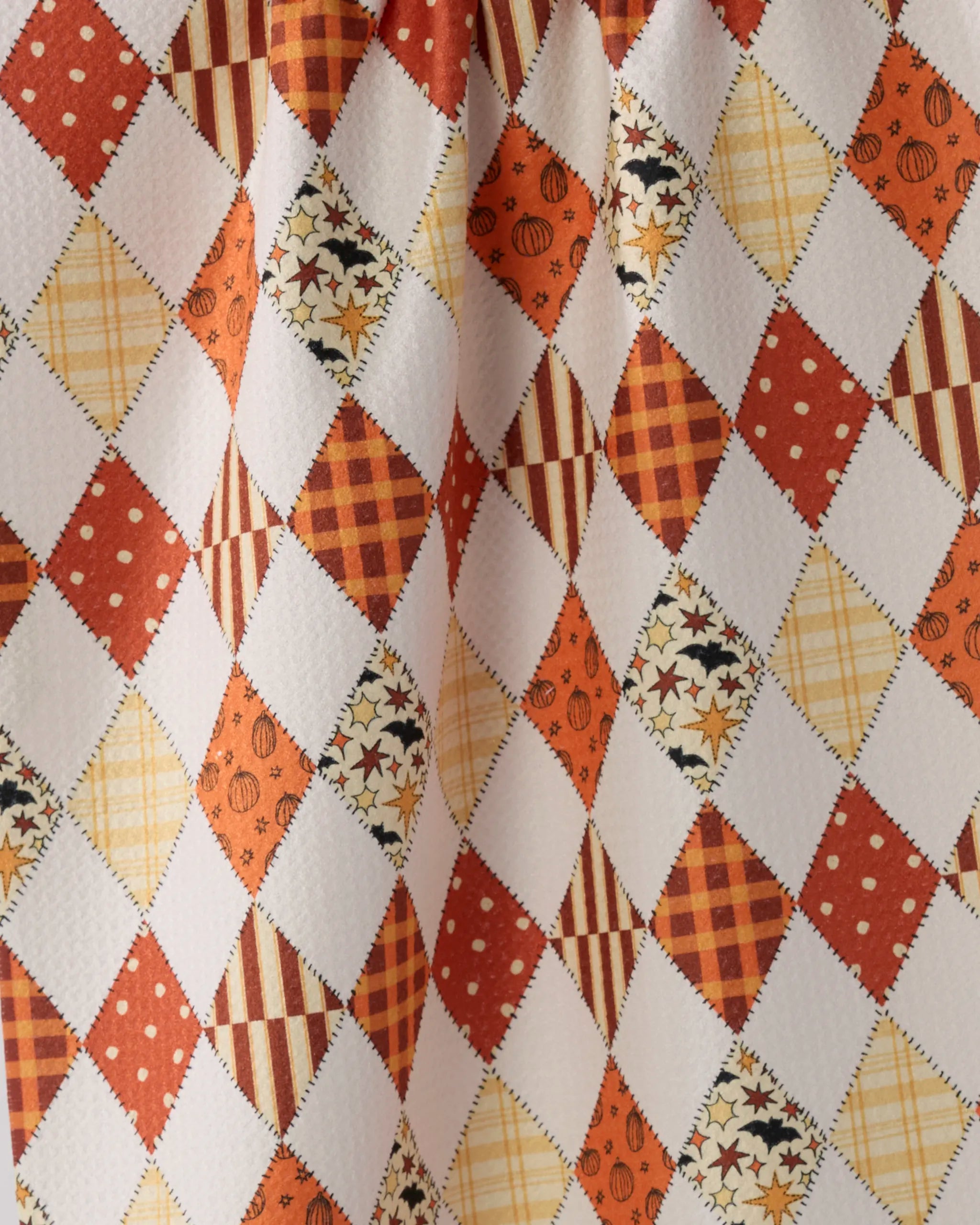 A close-up of the Geometry "Hayride" quick-drying towel displays an argyle pattern in orange, red, yellow, and cream with polka dots, stripes, plaid, and stars. The fabric is slightly wrinkled.