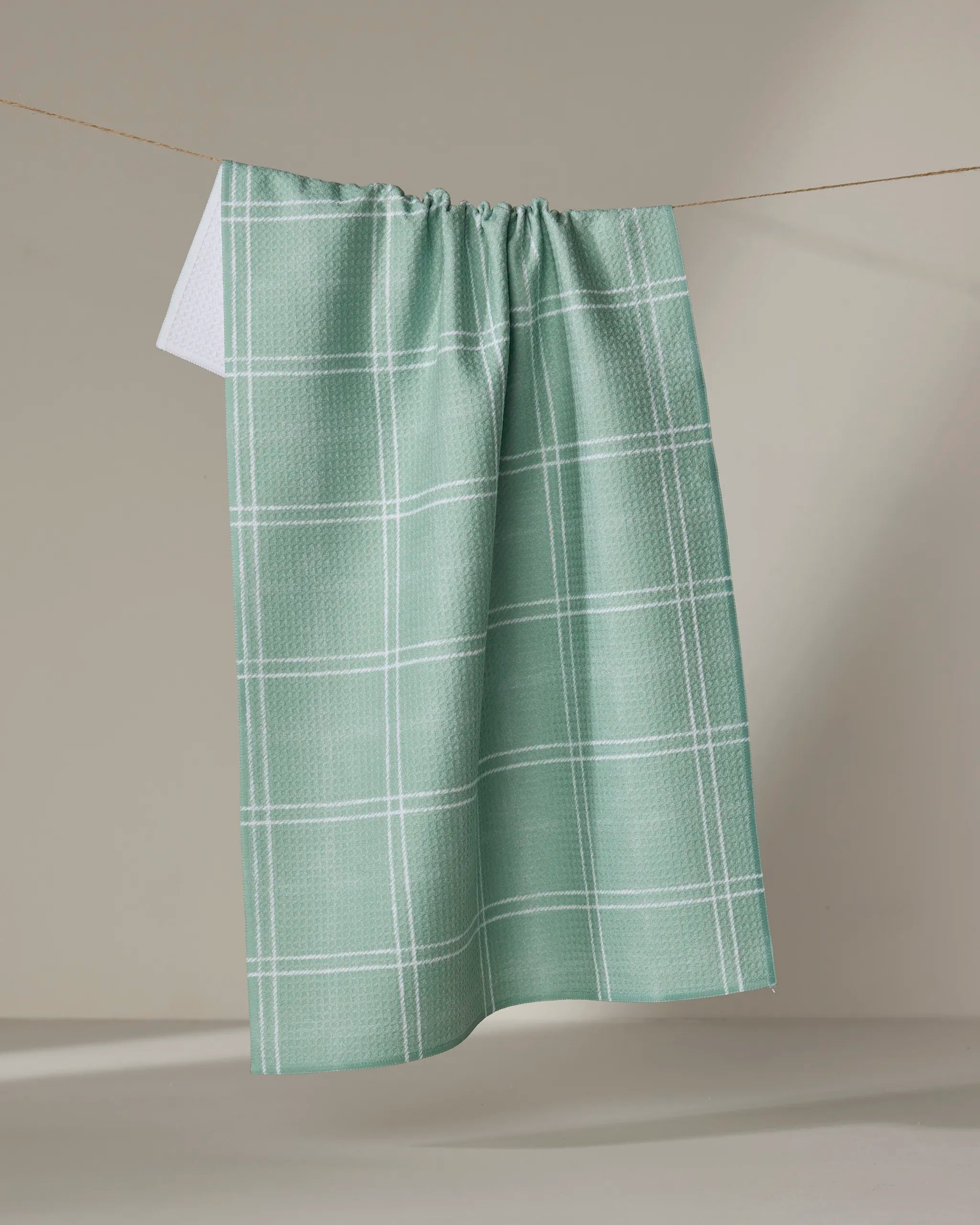 The Geometry Hearth Meadow dish towel, light green with a white checkered pattern, hangs on a clothesline against a neutral background.
