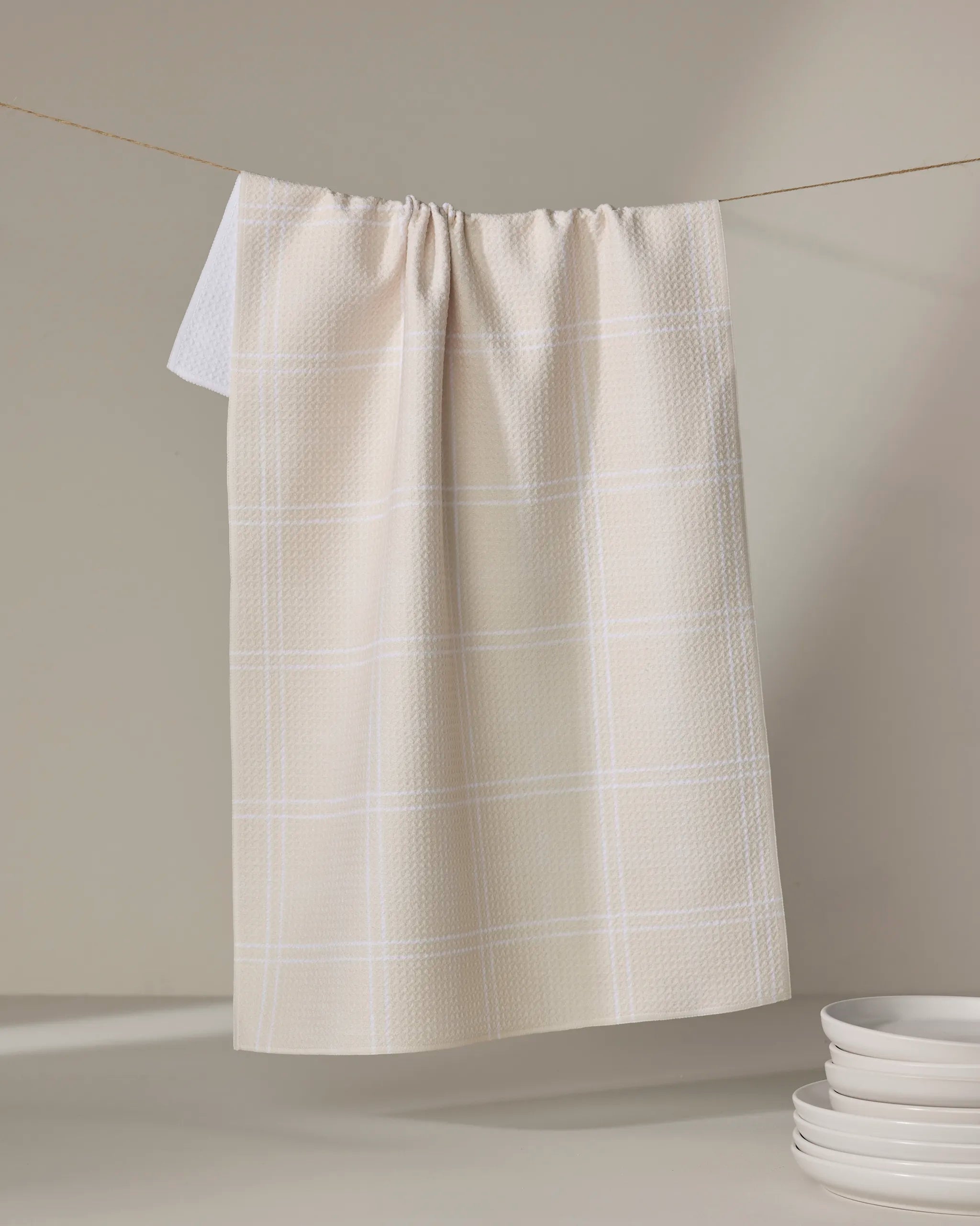 The Geometry Hearth Oat eco-friendly kitchen towel, featuring a beige and white checkered design, hangs on a string against a light background with a stack of white plates nearby.