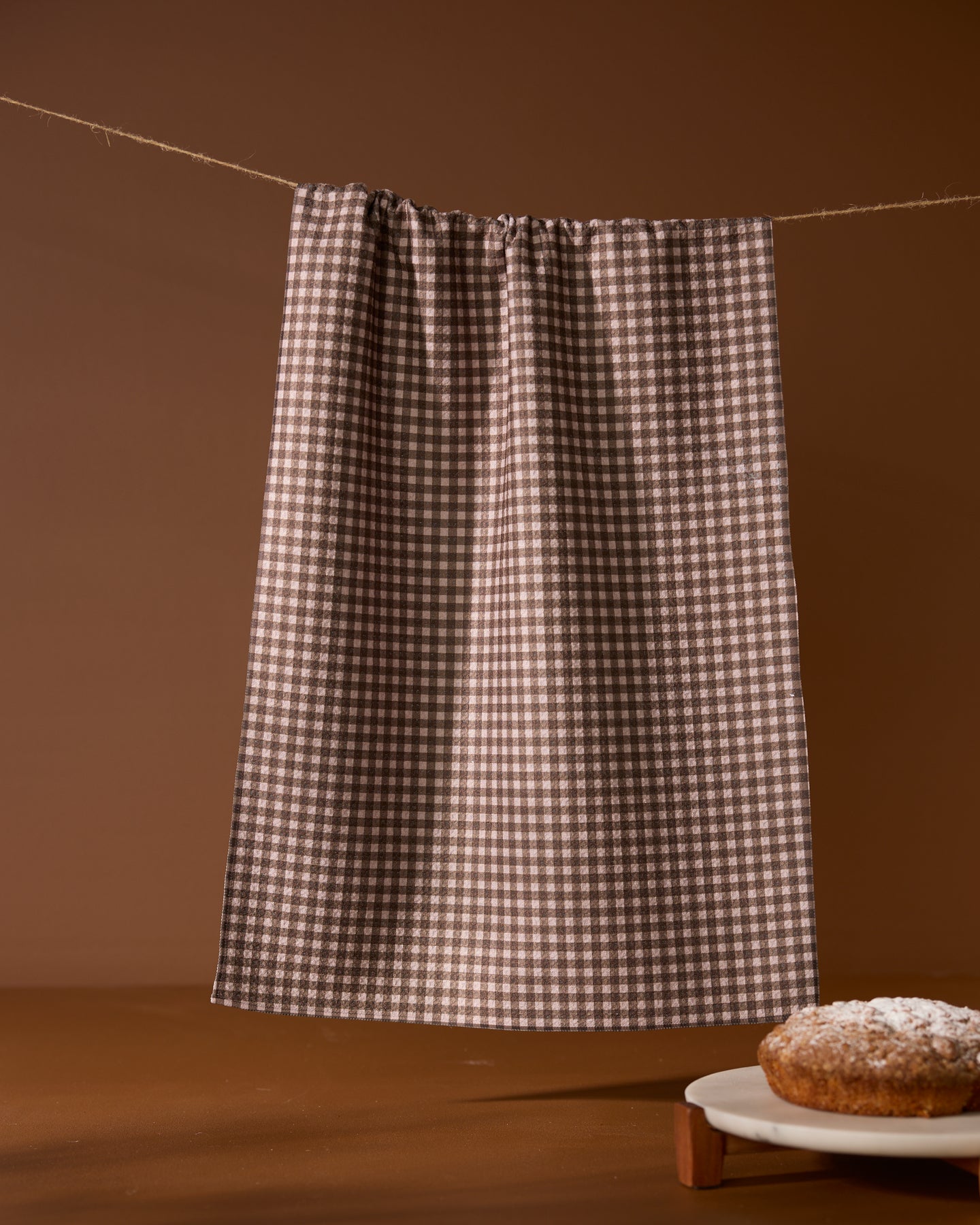A Geometry Hearth tea towel with a brown and white checkered pattern hangs on a string against a brown background. A powdered sugar-dusted cake sits partially visible on a wooden table in the bottom right corner.