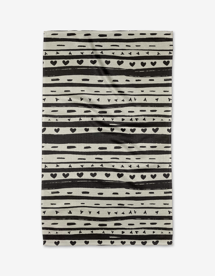 The Danelys Sidron Heartstring tea towel is rectangular and beige, made from recycled materials with black stripes, dots, hearts, and triangles in a repeating pattern. Slightly wrinkled, it is displayed on a white background.