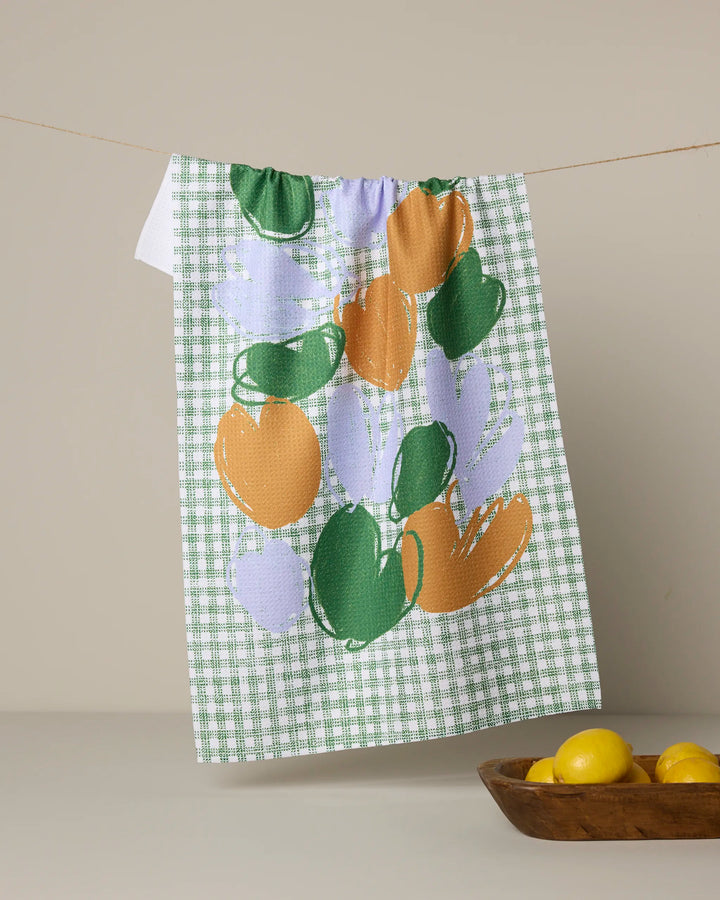 The Geometry Hej tea towel, with its green and white checks and abstract green, lavender, and orange shapes, hangs on a line. A wooden bowl of yellow lemons is in front. This quick-drying towel brings charm to any kitchen.