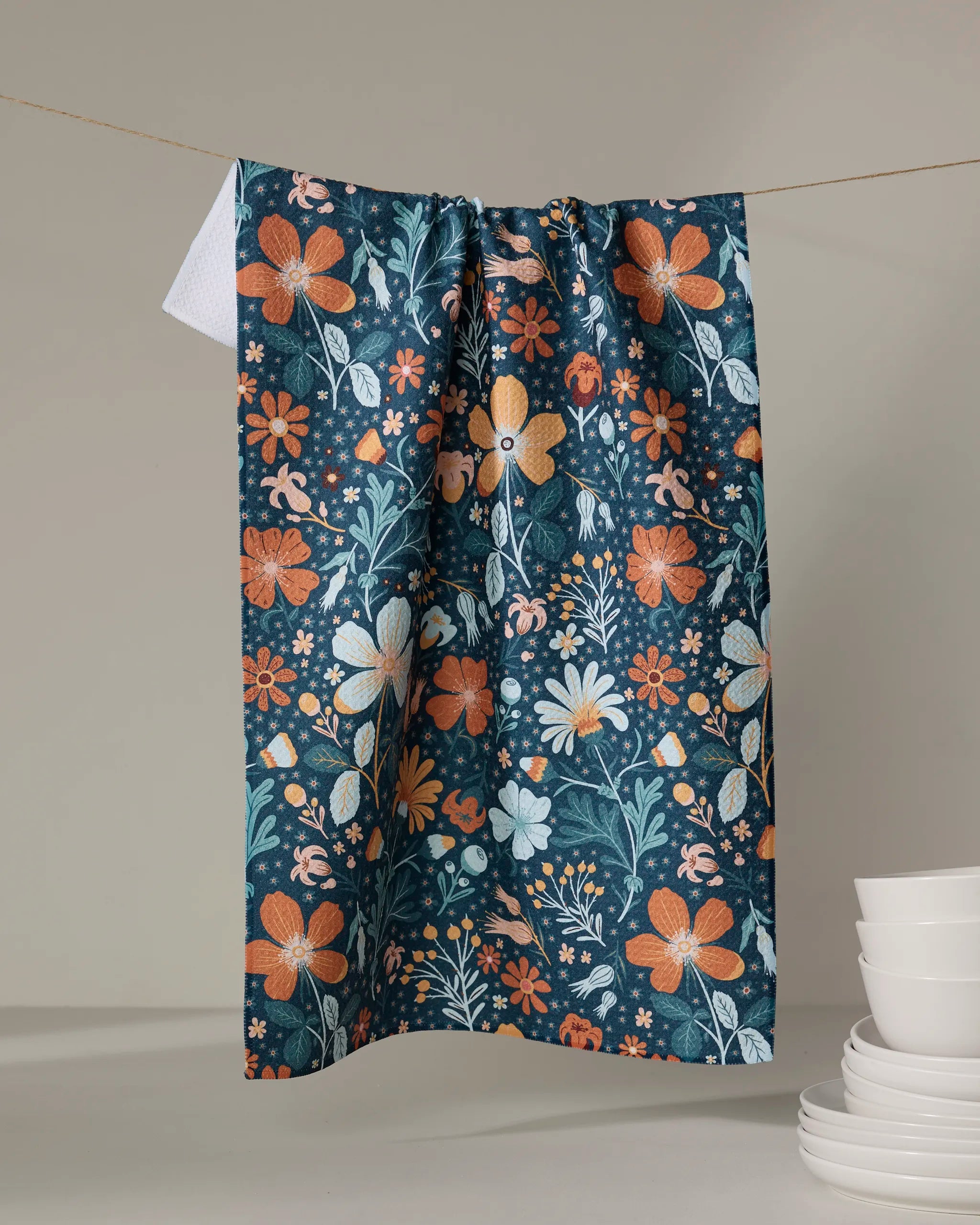 The Danelys Sidron Helle Bore tea towel, featuring an orange, yellow, and white floral design on dark blue, hangs on a line with white bowls stacked in the lower right corner against a neutral backdrop.