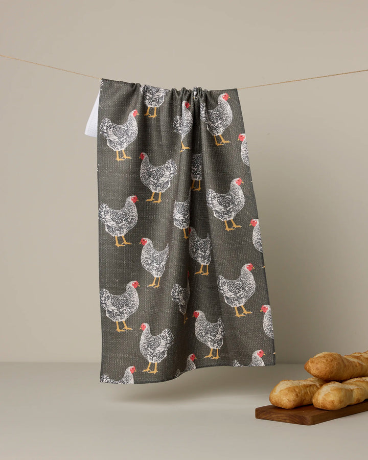 The Geometry Hen House gray kitchen tea towel, crafted from recycled materials and featuring white and black hens with red combs, hangs on a line. Three baguettes rest on a wooden board in front of a neutral beige background.