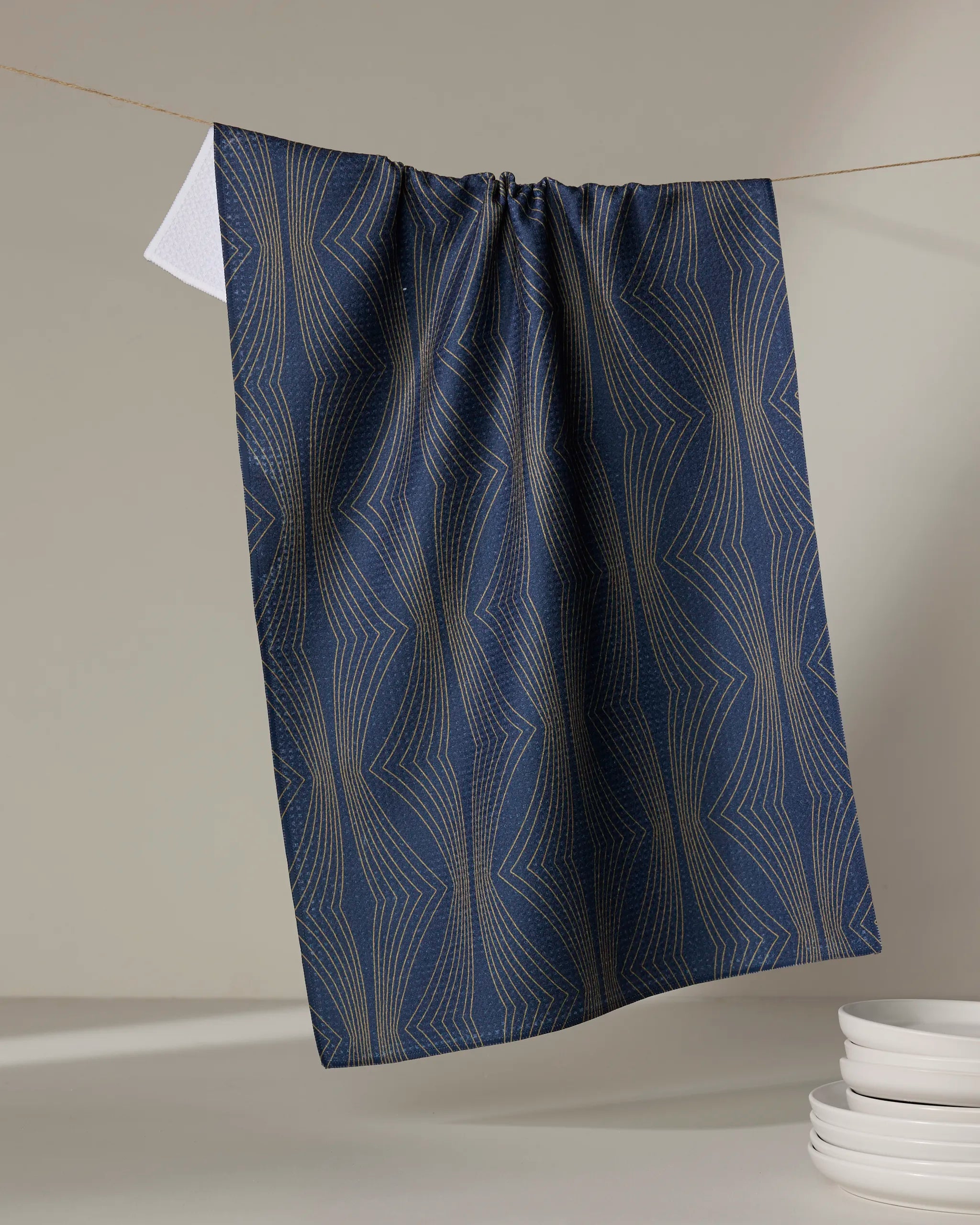 The Hera kitchen tea towel by Amy MacCready in dark blue with a gold geometric pattern hangs from a string, while stacked white plates are seen in the lower right corner against a neutral background.