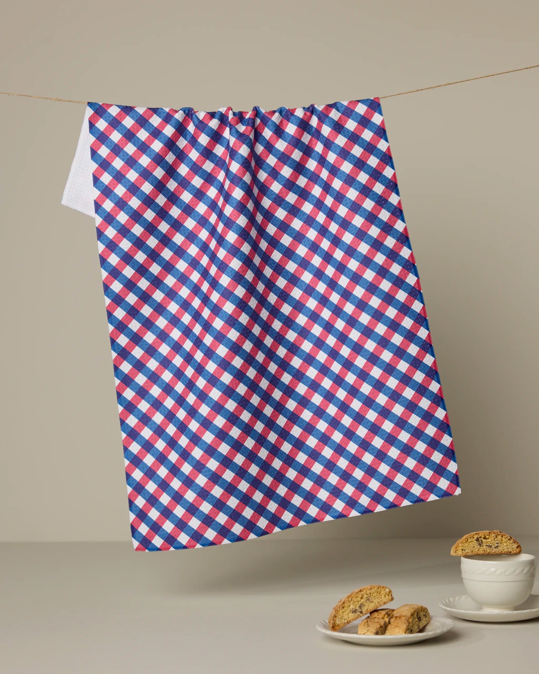 The Geometry Heritage Check ultra-absorbent tea towel in red, white, and blue hangs on a line against a beige background. Below, a white cup and plate with biscotti sit on a light gray surface.