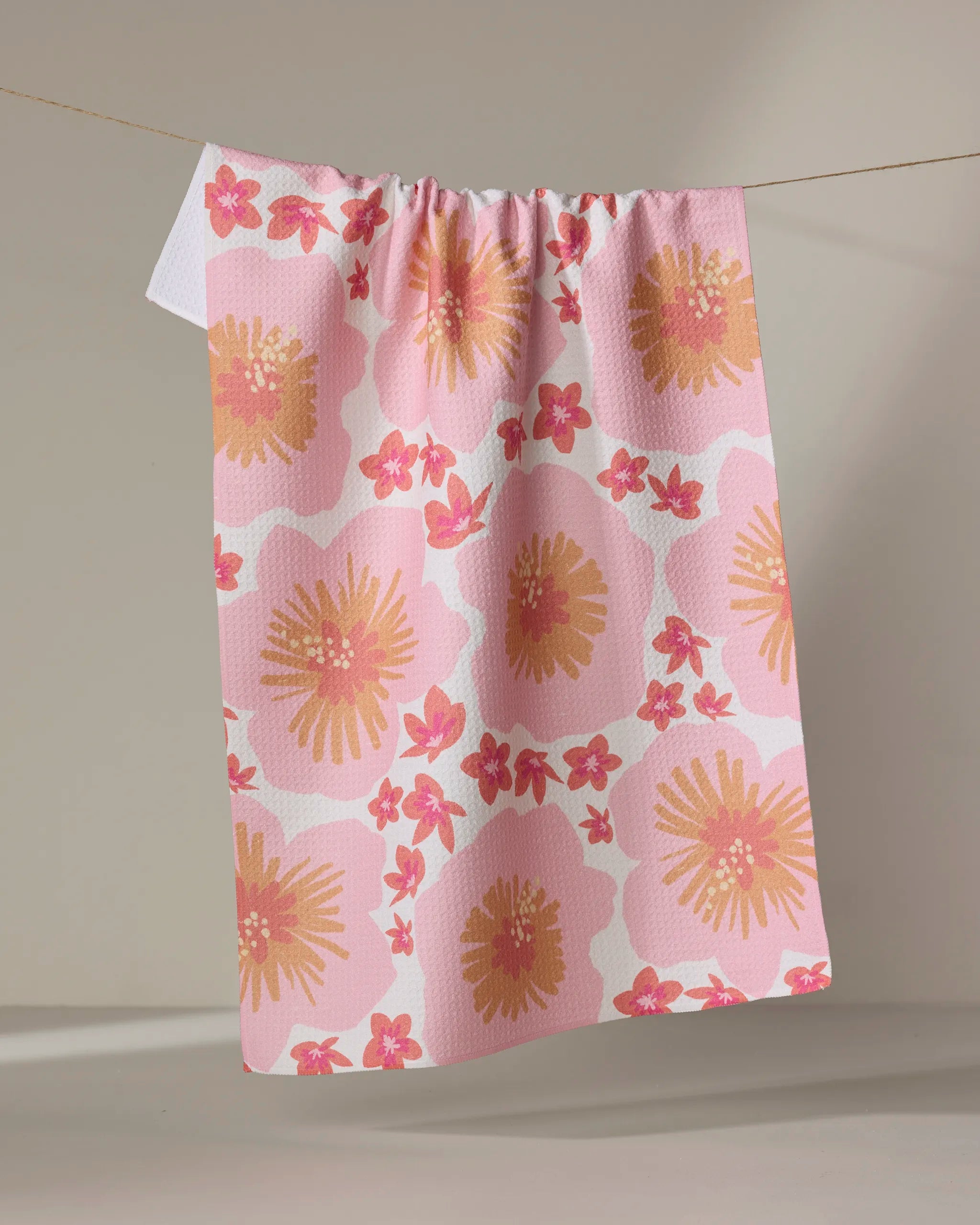 The Hibiscus Sunset kitchen towel by Janna Sue Design, made from recycled materials and featuring vibrant floral patterns in pink, yellow, and orange, hangs on a clothesline against a neutral background.