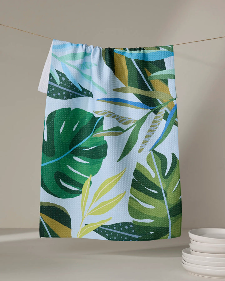The Hidden Island kitchen tea towel by Janna Sue Design, featuring a vibrant green and yellow tropical leaf pattern, hangs on a line with a neutral background and white plates stacked in the lower right corner.