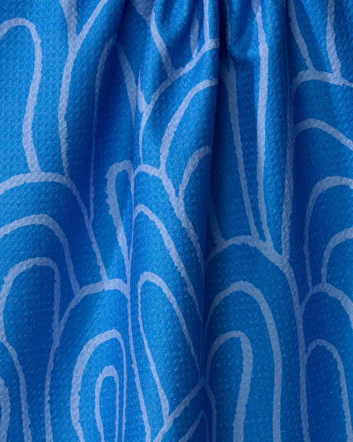 A close-up of the Sareka Unique "High Tide" tea towel shows blue recycled fabric with a light blue wavy pattern, gathered ruching at the top, and a slightly pebbled texture.