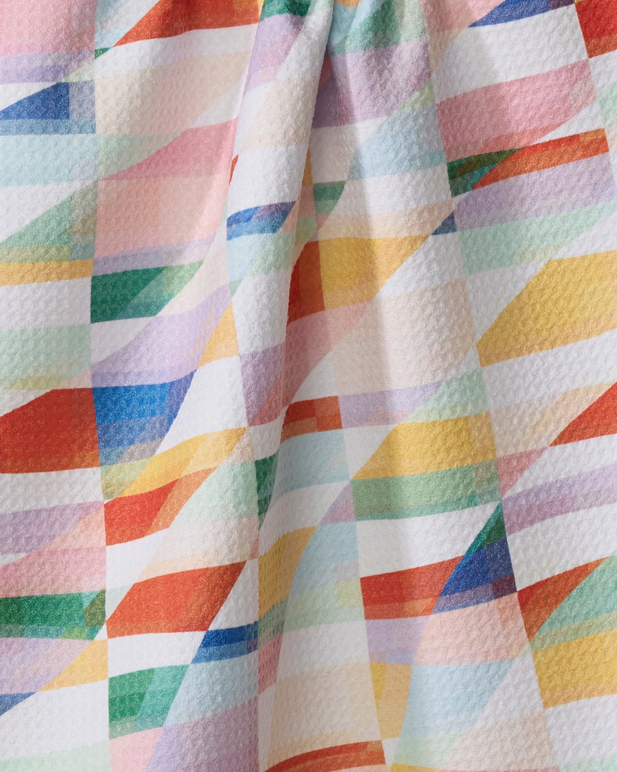A close-up of the Rachel Parker "Highlands" dish towel, featuring a colorful geometric pattern with intersecting lines in red, blue, yellow, green, and purple on a textured white background.