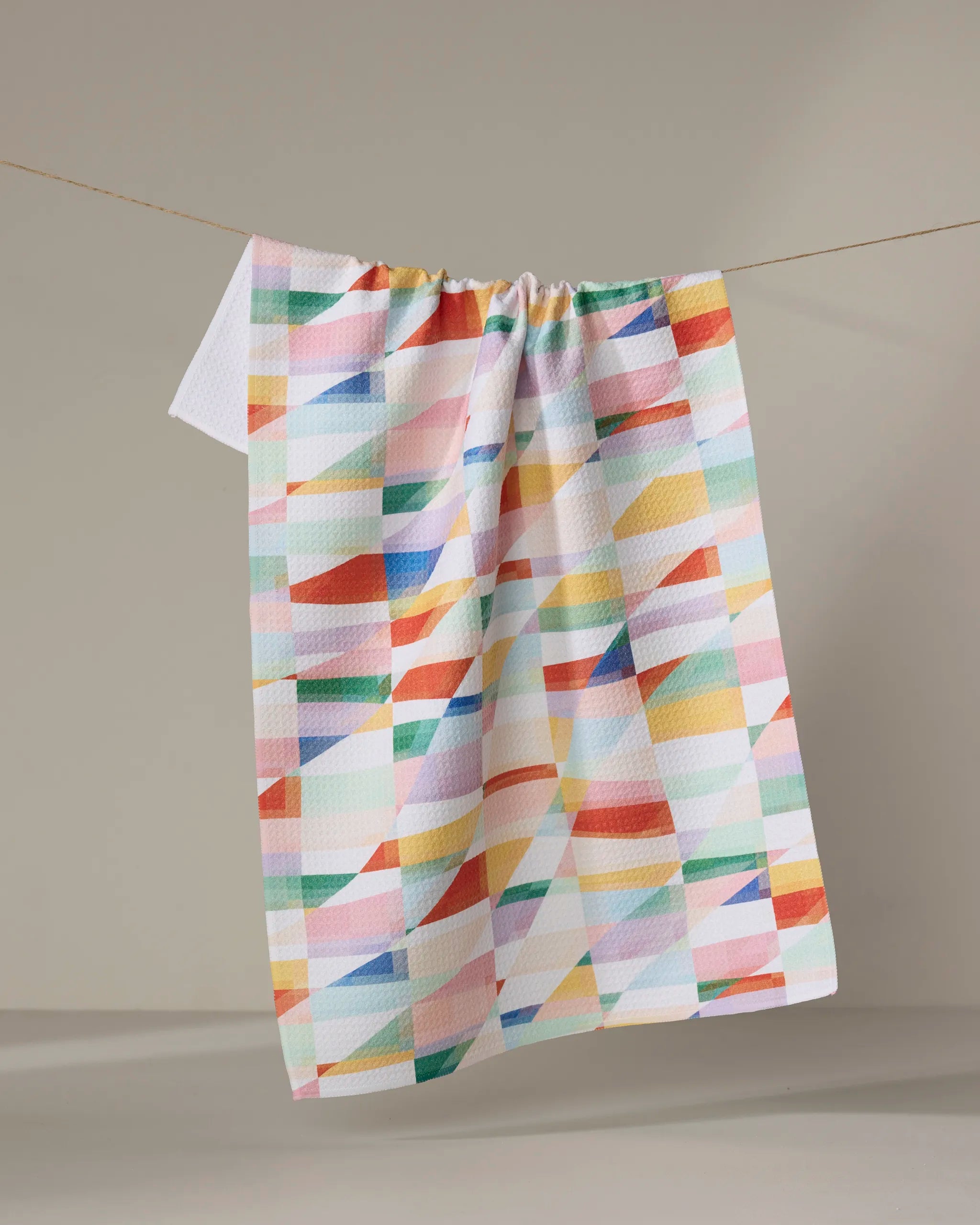The "Highlands" recycled kitchen towel by Rachel Parker features a vibrant geometric abstract pattern in red, blue, yellow, orange, and green and hangs on a line against a neutral background.
