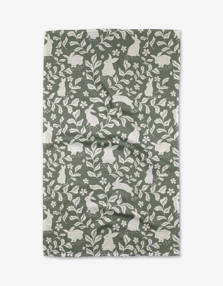 The Hippity Hop tea towel by Charlie Rowan Designs features a repeating pattern of white rabbits, leaves, and flowers in green and white on slightly wrinkled recycled fabric, laid flat on a white background.
