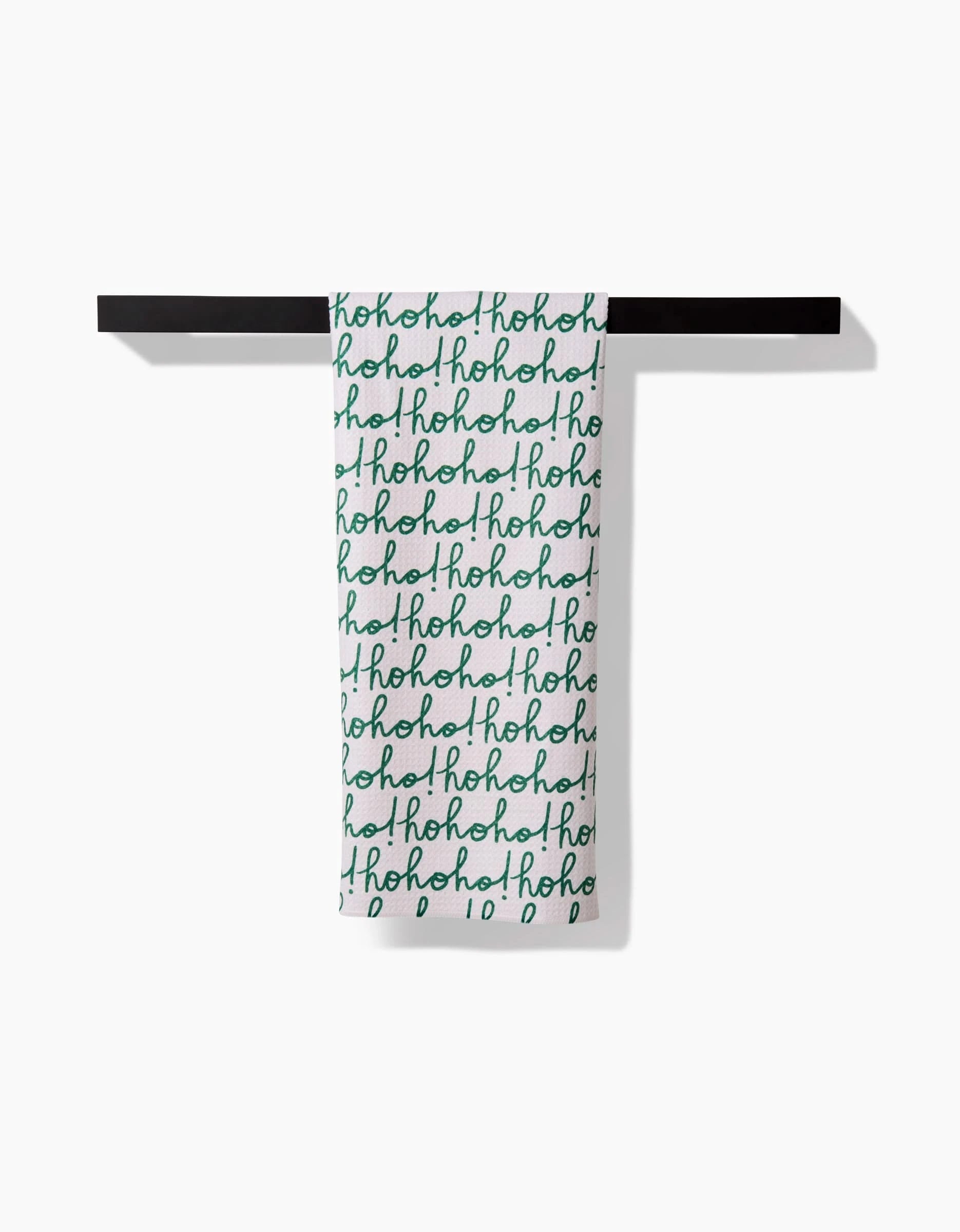 The Kayla Ann Ho Ho Holiday Green white tea towel hangs on a black bar, featuring festive green handwritten ho! ho! ho! text in a repeated pattern.
