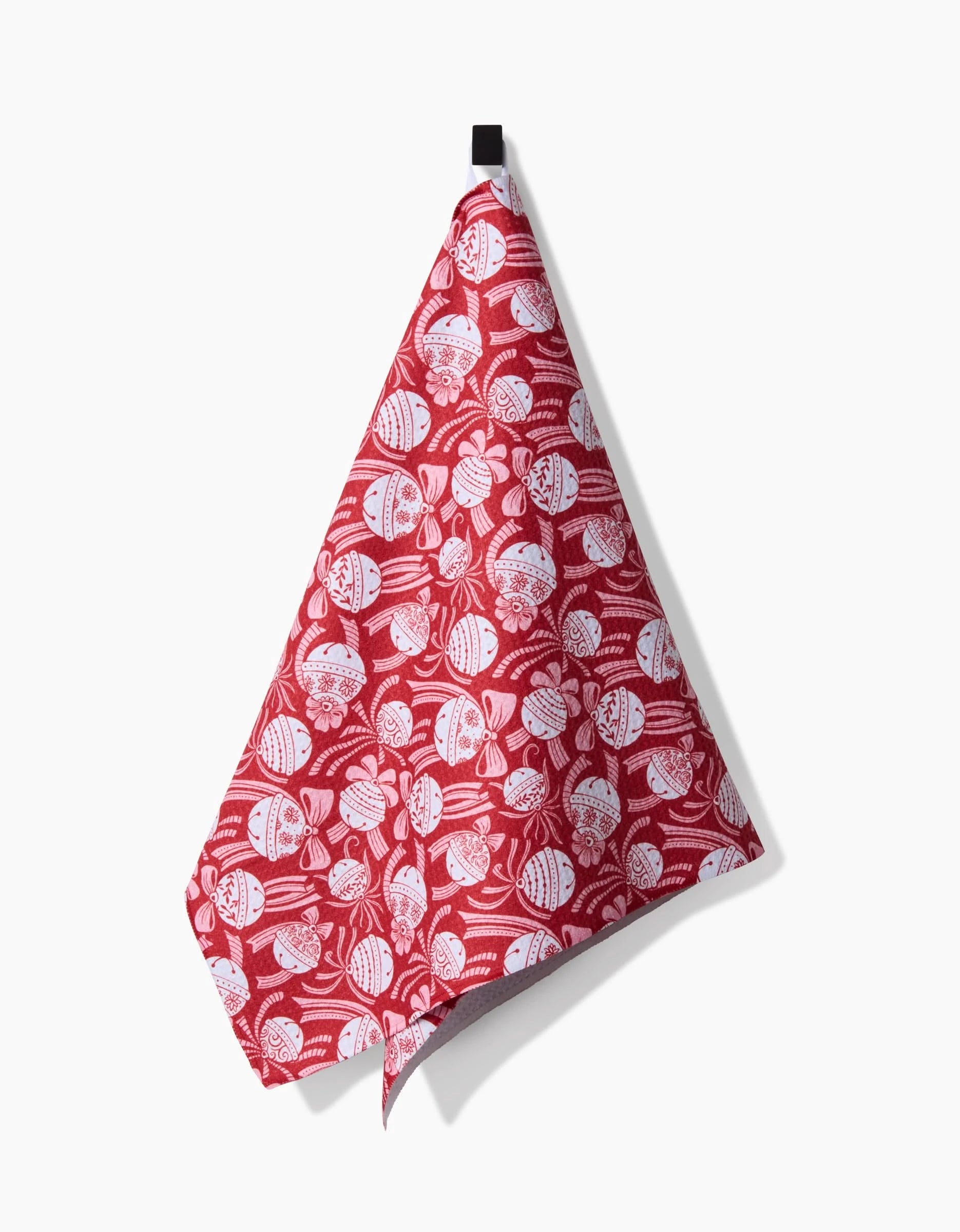 The Holiday Bells Bows tea towel by Sarah Daugherty, featuring a festive white pattern on red and made from recycled materials, hangs from a black hook against a white background.
