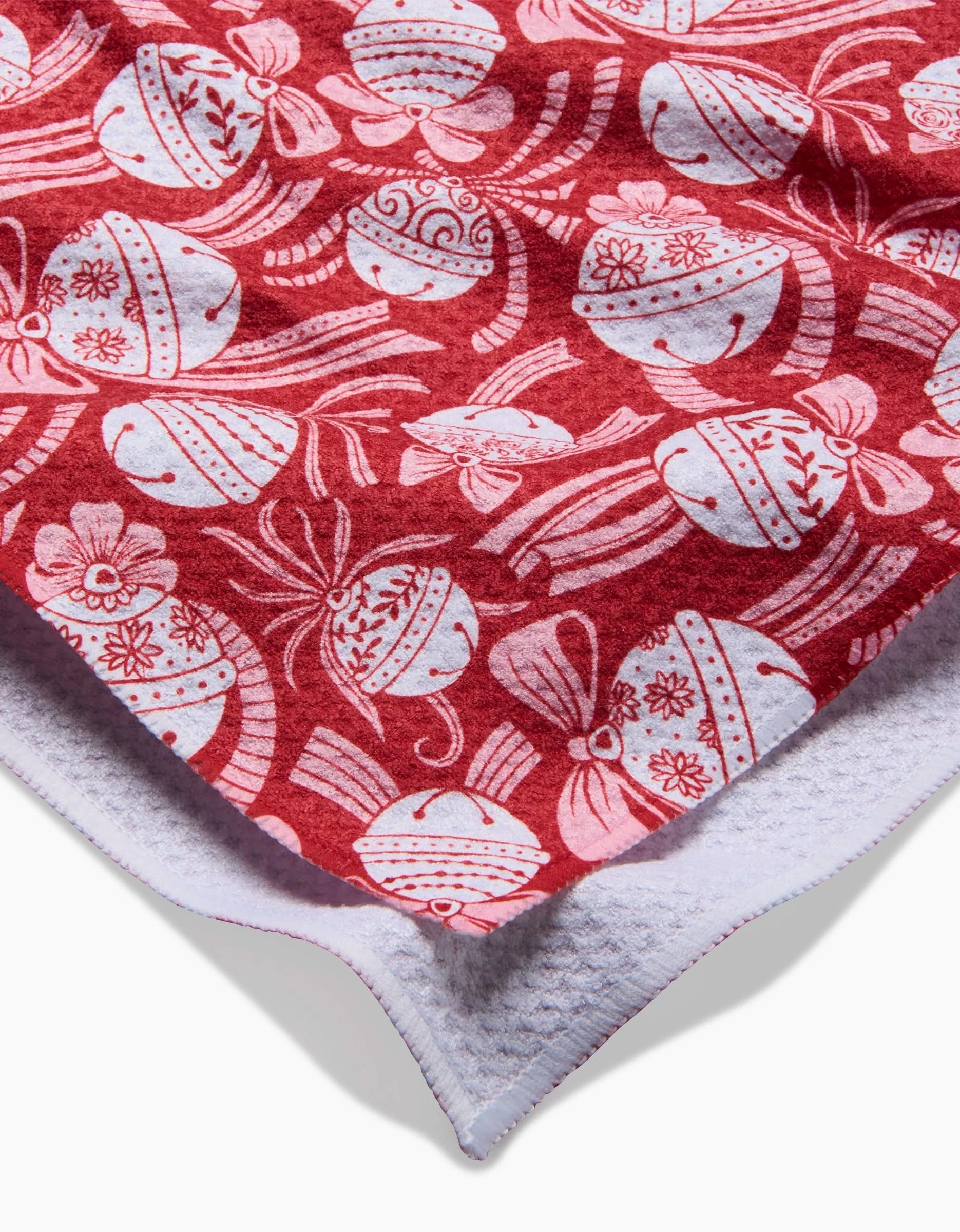 A close-up of the Sarah Daugherty Holiday Bells Bows tea towel in red and white, showing ornaments, bows, and flowers on festive fabric with a quick-drying white textured back.