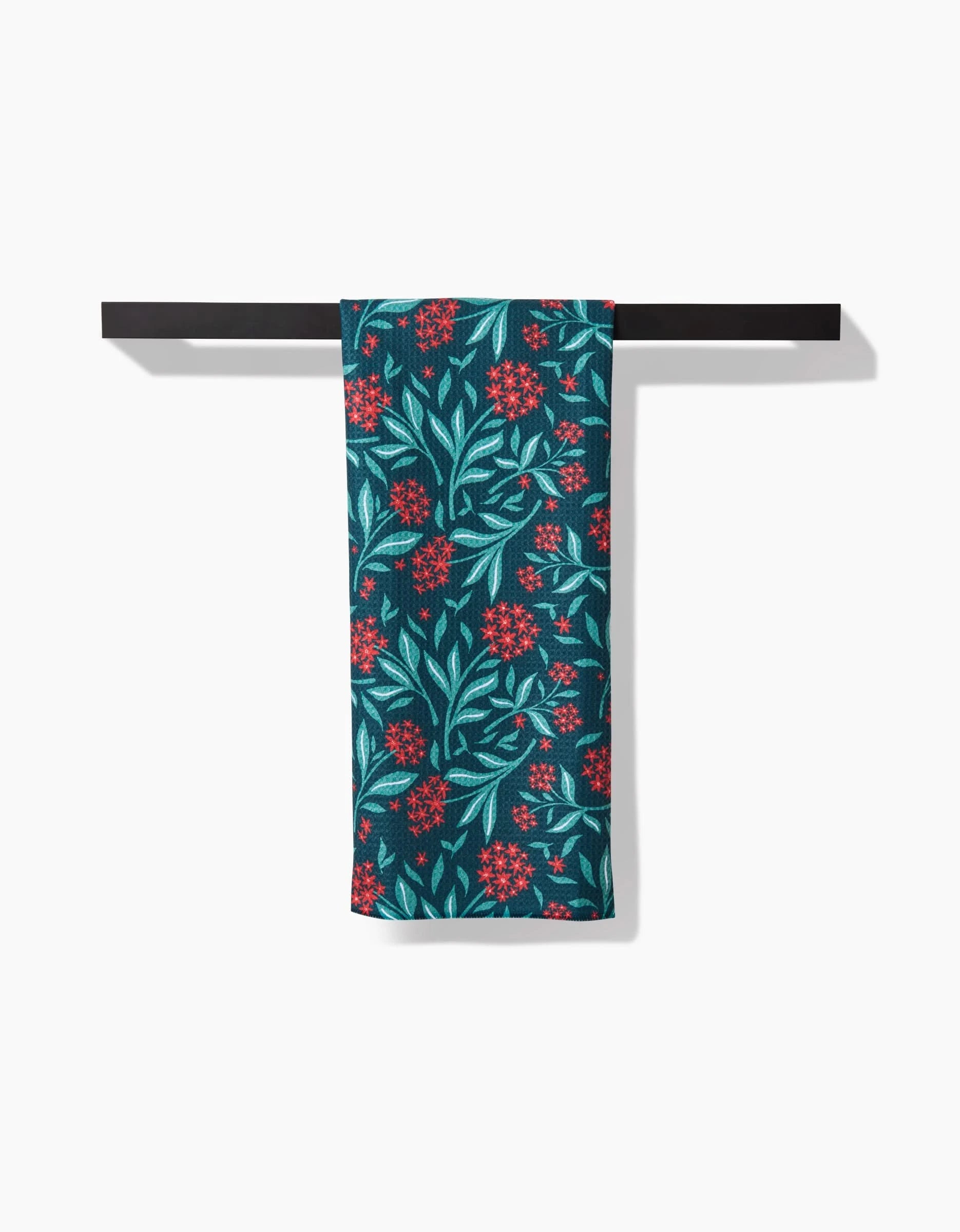 The Holiday Blooms tea towel by Charlie Rowan Designs, featuring a red and light blue floral pattern on dark teal and made from recycled materials, hangs neatly on a black towel bar against a white background.