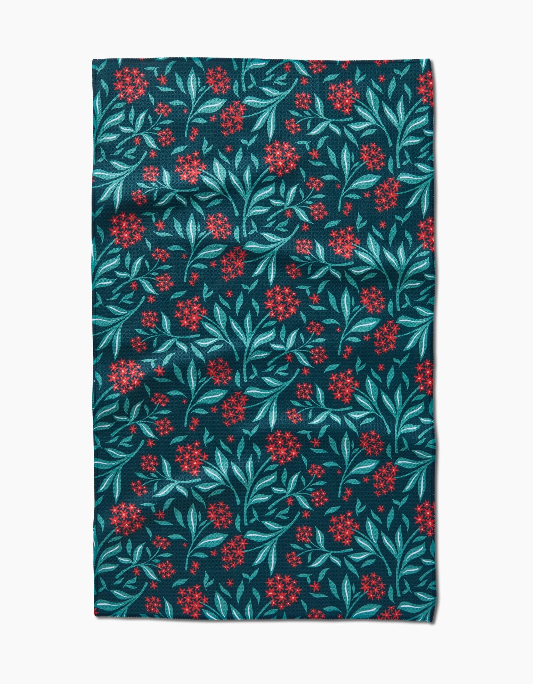 The “Holiday Blooms” tea towel by Charlie Rowan Designs features a dark background with teal leaves and red flowers, crafted from quick-drying recycled materials.