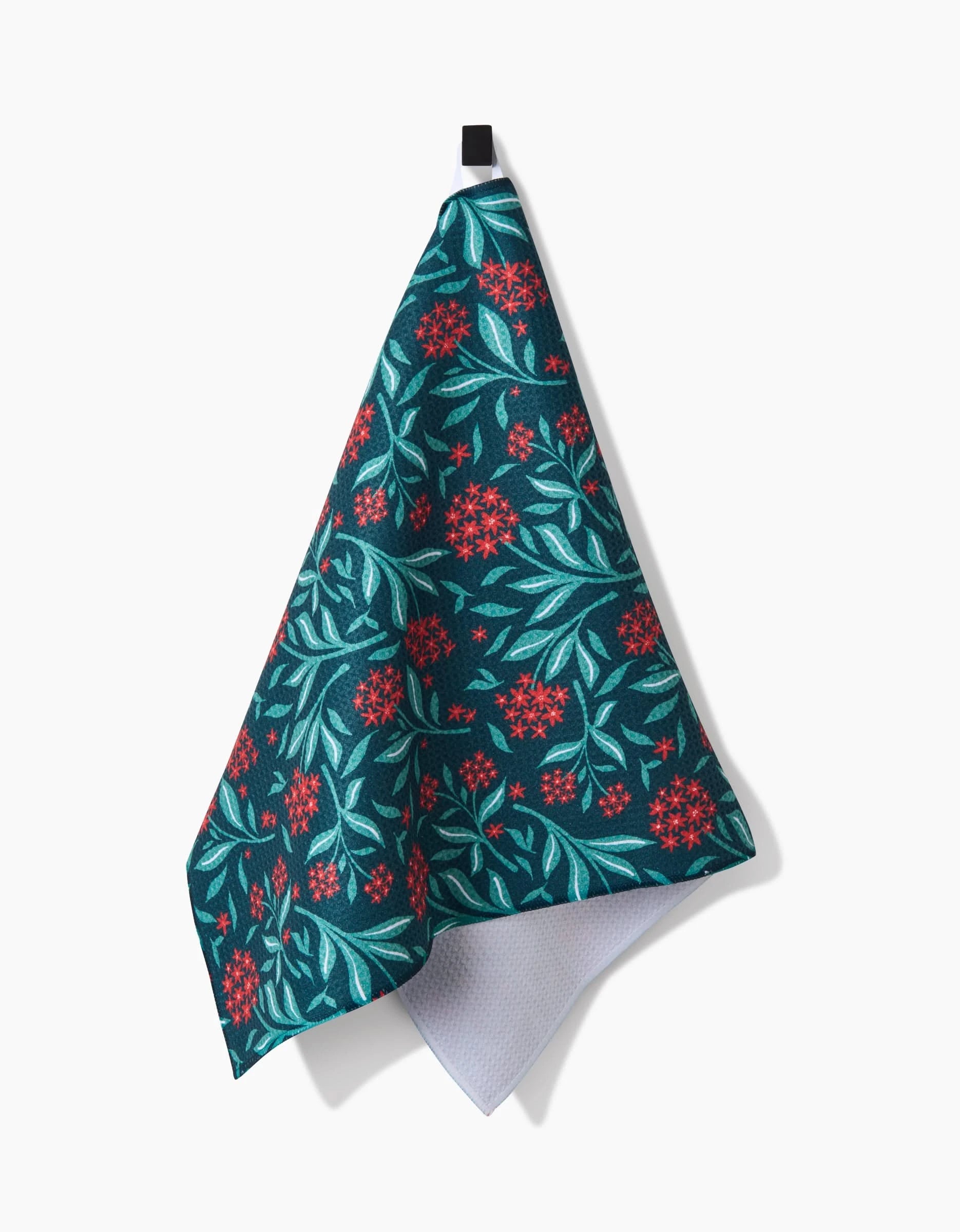 The Holiday Blooms tea towel by Charlie Rowan Designs features red flowers and light green leaves on dark green, a black hanging loop, and a turned-up corner showing the white underside. Made from quick-drying recycled materials.