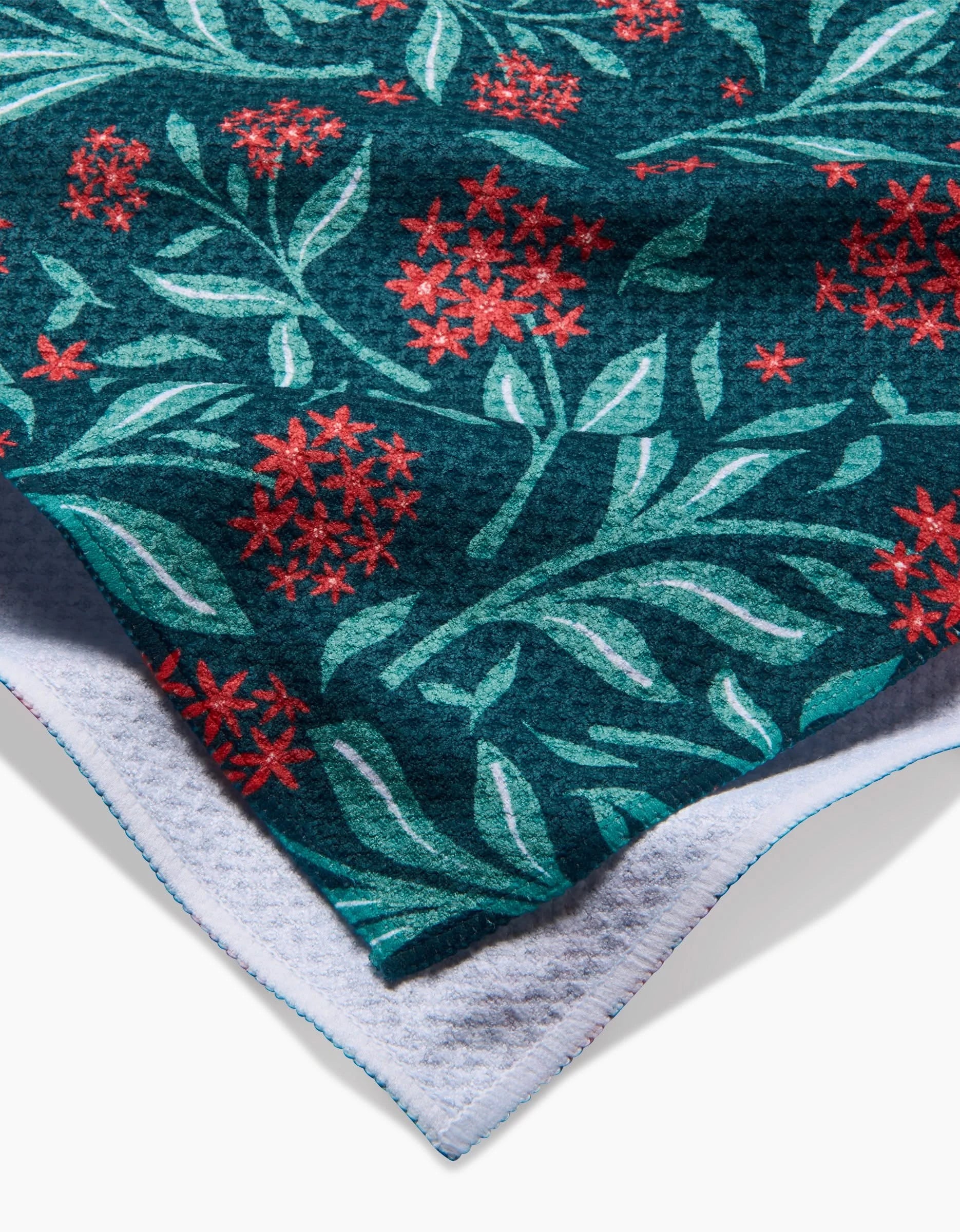 A close-up of the Holiday Blooms tea towel by Charlie Rowan Designs, featuring a red and green floral pattern on dark teal, with white lining underneath and blue edge stitching.