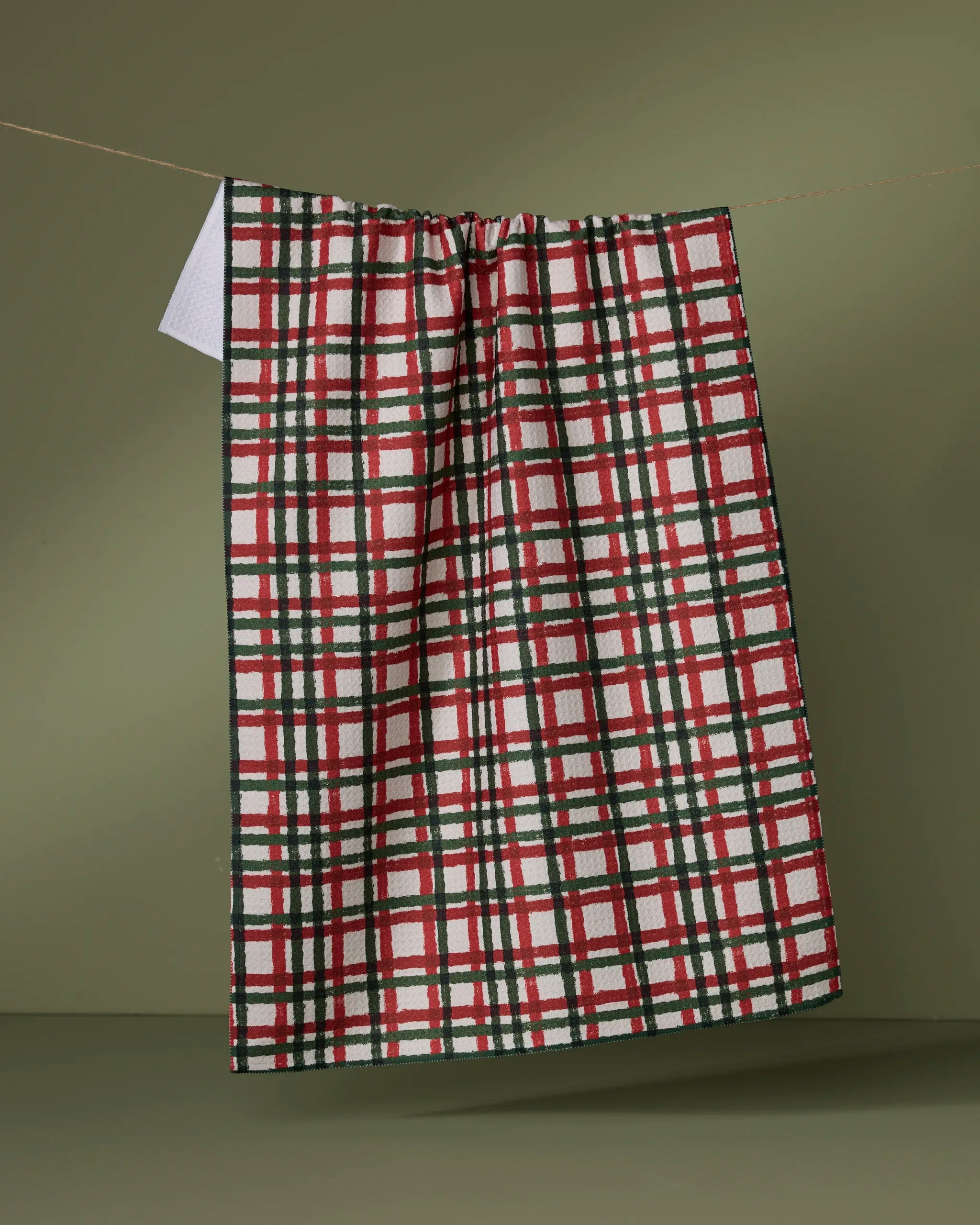 The Geometry Holiday Charm tea towel, featuring an ultra-absorbent red, green, and white plaid design, hangs on a string against an olive green background.