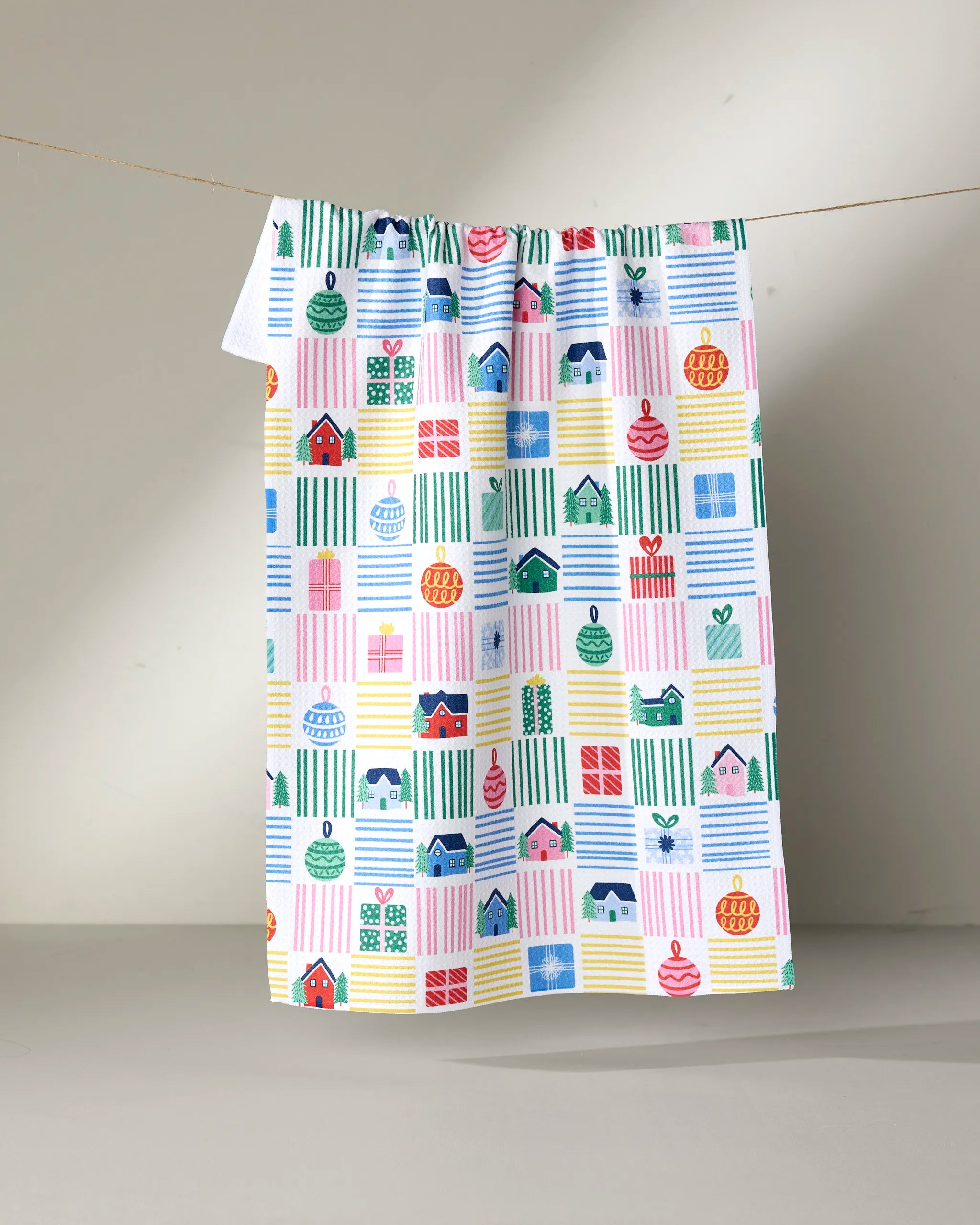 The Holiday Checkered tea towel by Ceyda Alasar, made from recycled materials, features pastel and bright colors in a playful grid of whimsical houses, plants, and shapes. It hangs on a string against a plain light gray background.