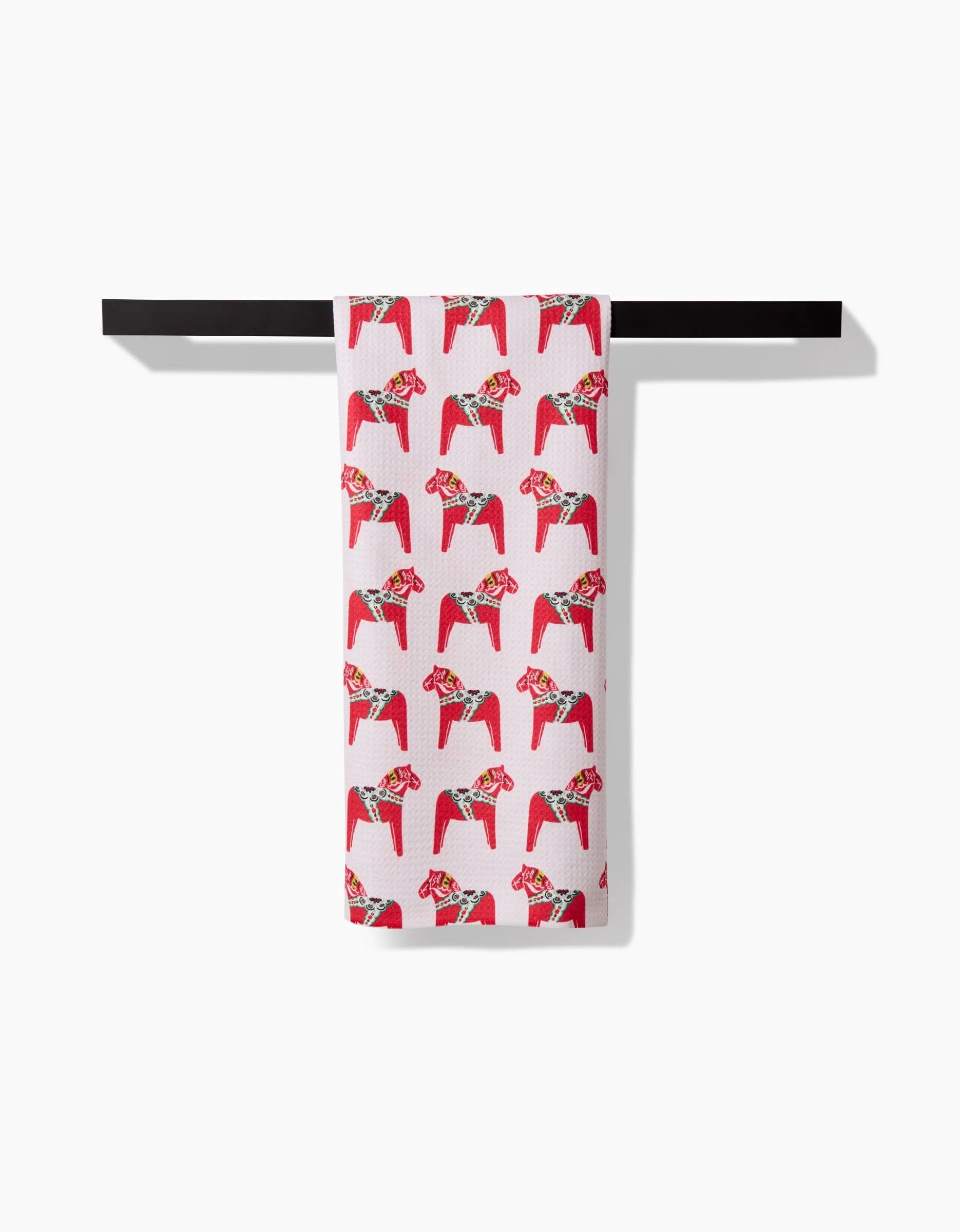 A quick-drying white Holiday Dala Horse tea towel by Kayla Ann, featuring a pattern of red and pink dala horses with floral crowns, hangs neatly on a black towel bar against a white background.