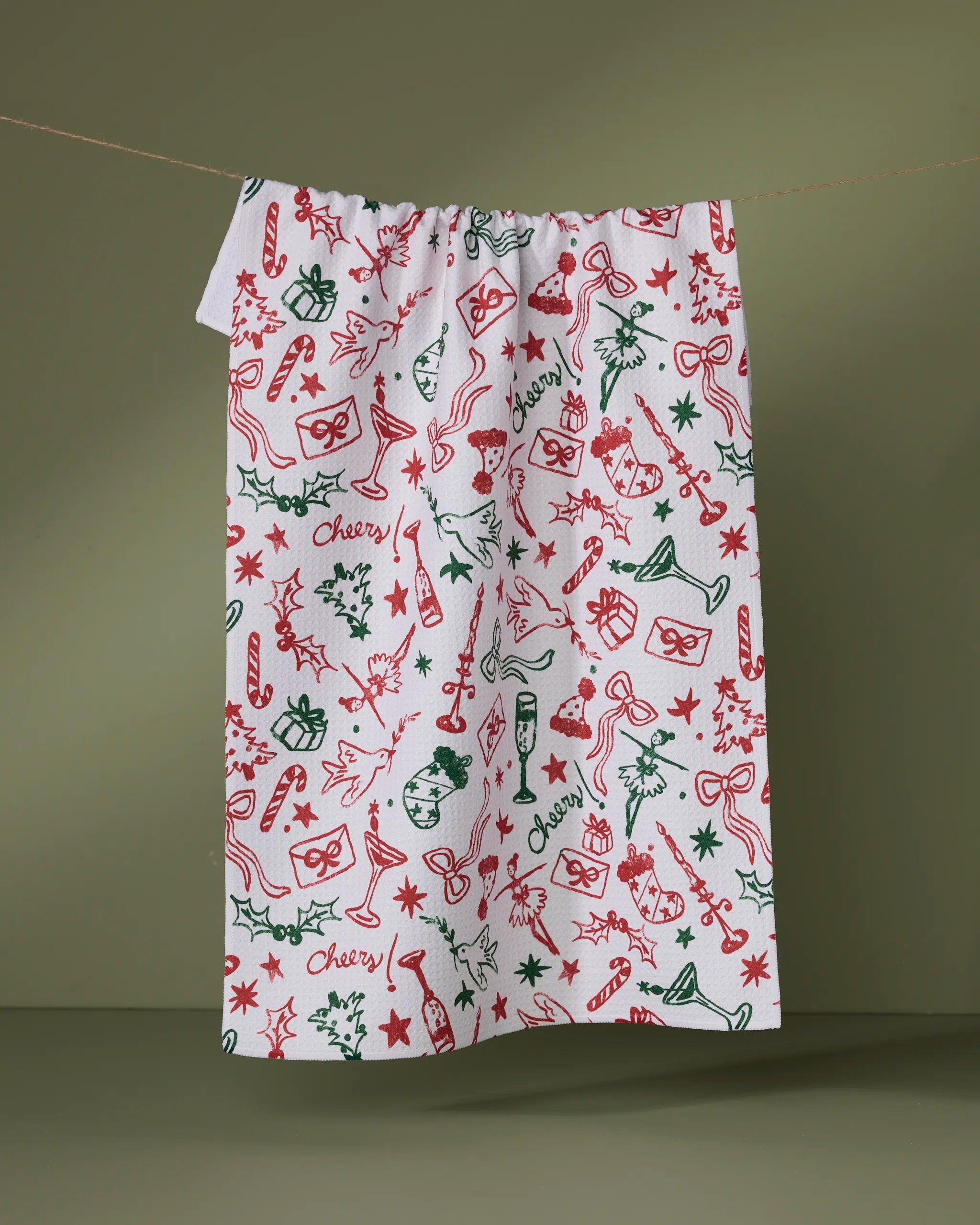 The "Holiday Doodles Multi" tea towel by Geometry is made from recycled materials and features festive red and green illustrations—candy canes, stars, presents, champagne glasses, bows, and "cheers"—against a green background.