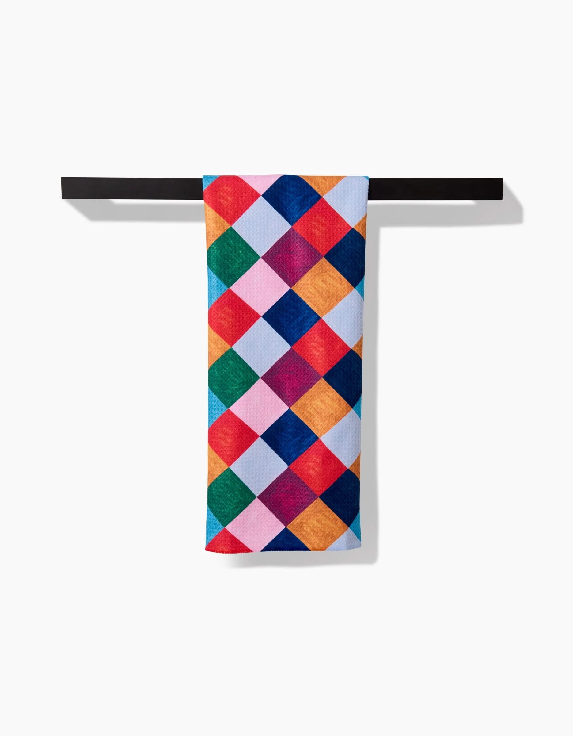 The Holiday Harlequin tea towel by Daria Solak features a vibrant diamond pattern and hangs neatly over a simple black rod against a white background.