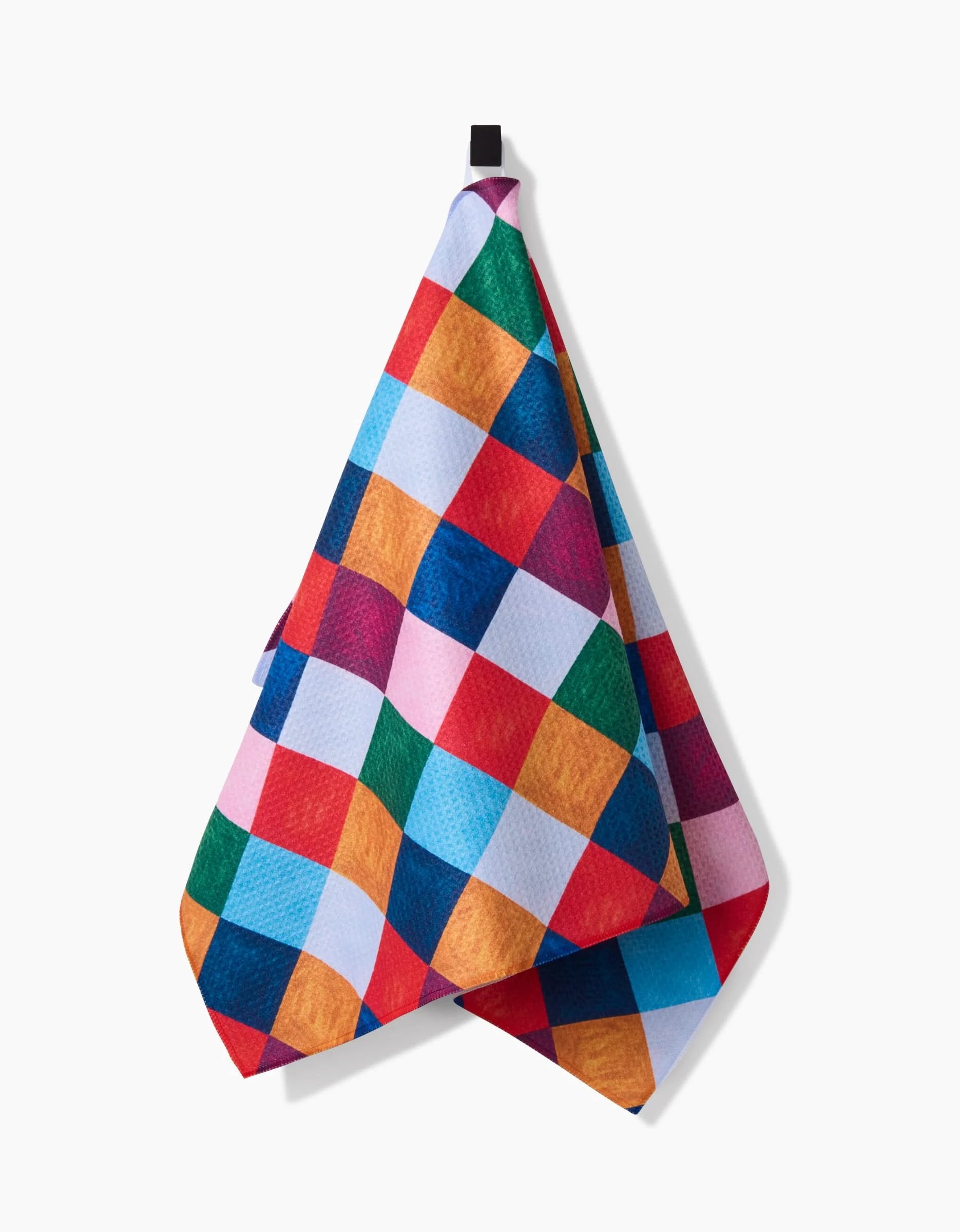 The Daria Solak Holiday Harlequin kitchen towel, featuring a vibrant checkered pattern in red, blue, yellow, green, pink, and white squares, hangs from a black hook against a plain white background.