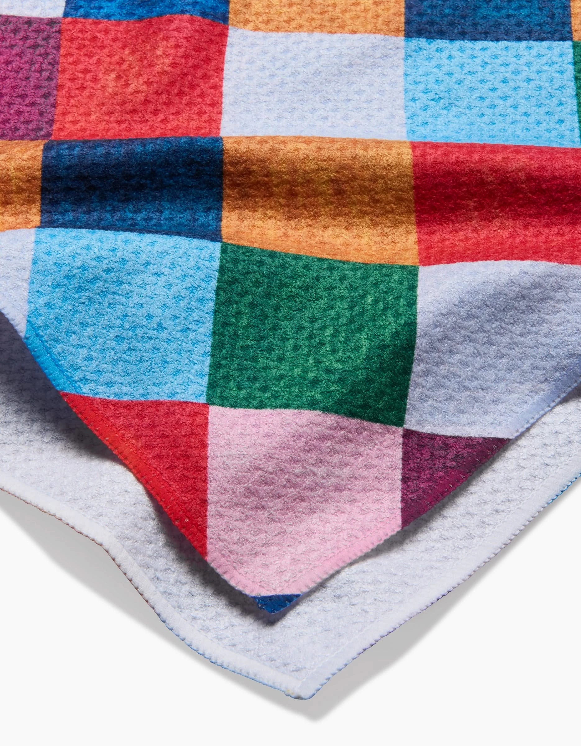A close-up of the Holiday Harlequin blanket by Daria Solak, showing its colorful checkered pattern (red, blue, green, yellow, pink) and light gray underside—crafted from recycled materials for eco-friendly comfort.