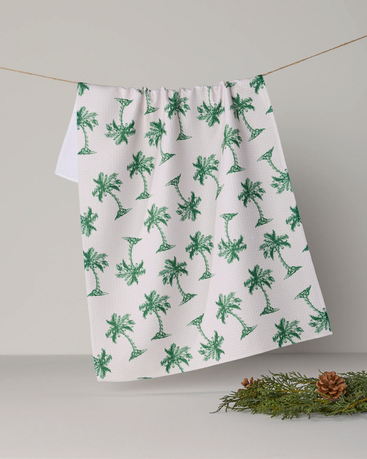 The Geometry “Holiday Hideaway” recycled kitchen towel, featuring a green palm tree pattern, hangs on a string against a light gray wall, with a pinecone and greenery arranged on the surface below.