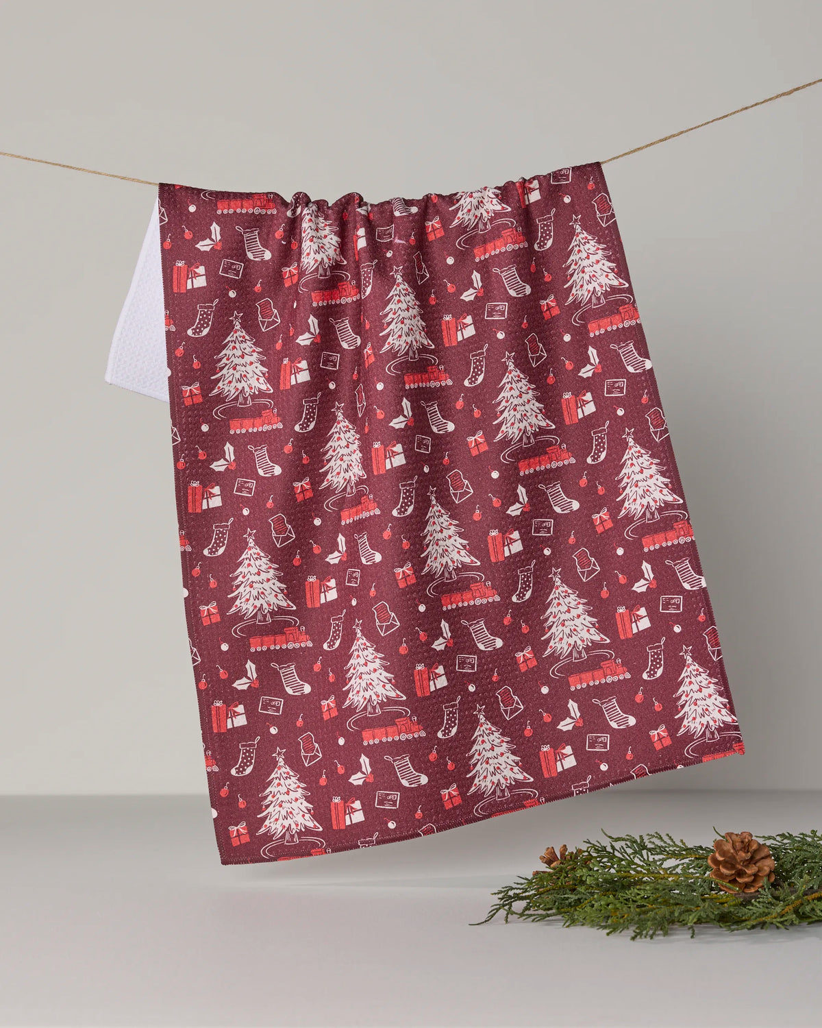 The Geometry Holiday Jubilee tea towel, crafted from recycled materials, features a festive red background adorned with white Christmas trees, stockings, and gift patterns. Hanging on a line above pinecones and greenery, this charming piece adds cheerful holiday style to any space.