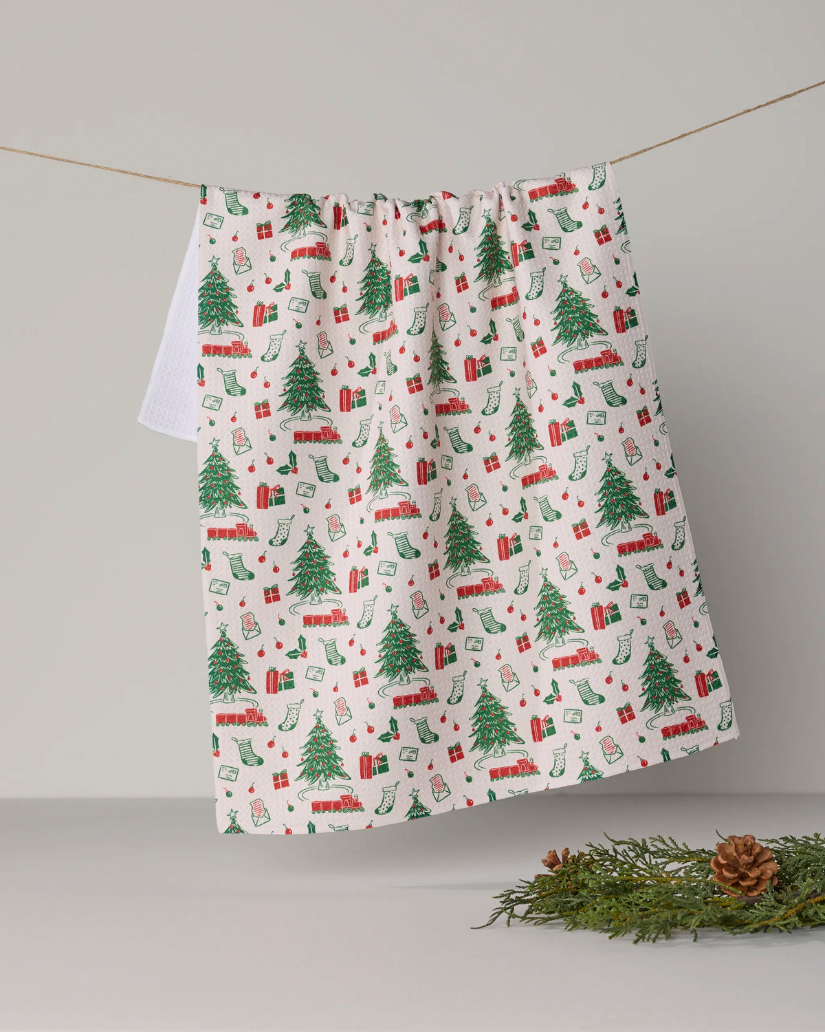 The “Holiday Jubilee” tea towel by Geometry features festive patterns of Christmas trees, gifts, stockings, and trains hanging on a line. Made from durable Geoweave fabric, this towel blends cheerful holiday design with practicality. Placed below on a gray surface, a small pine branch with pinecones enhances the seasonal charm.