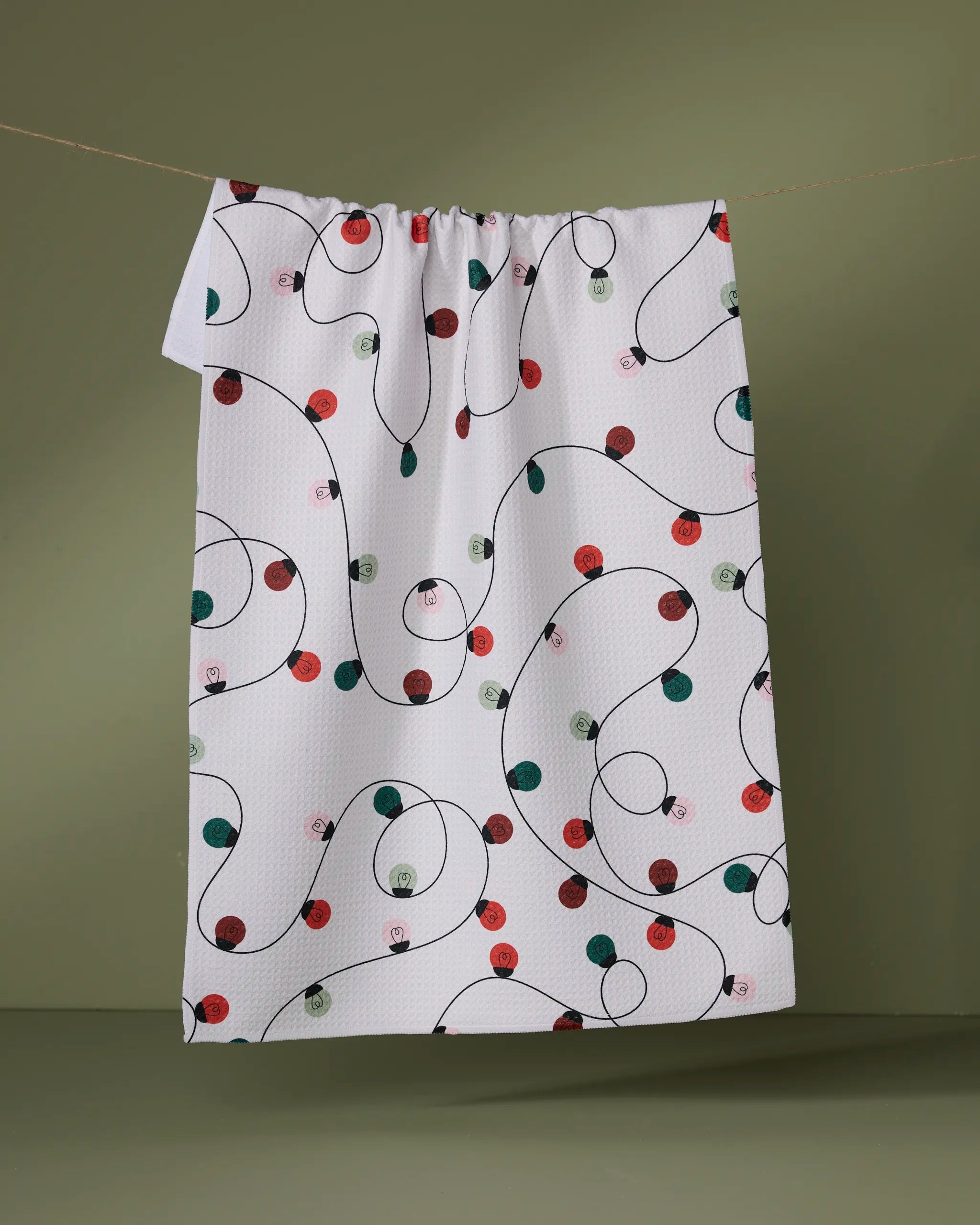 The Geometry “Holiday Lights” towel, ultra-absorbent and white with black winding lines plus red, green, and pink dots, hangs on a line against a muted olive green background.